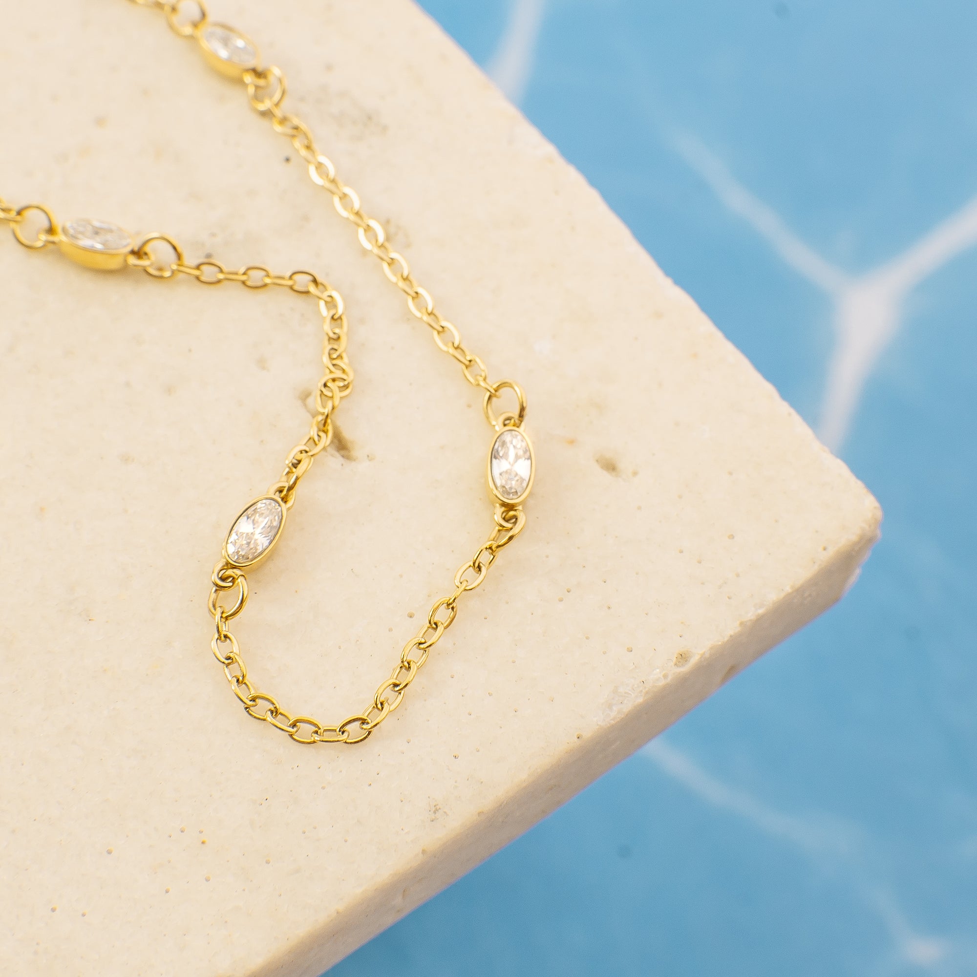 Gold chain necklace with clear stones on a beige stone surface with a blue tiled background