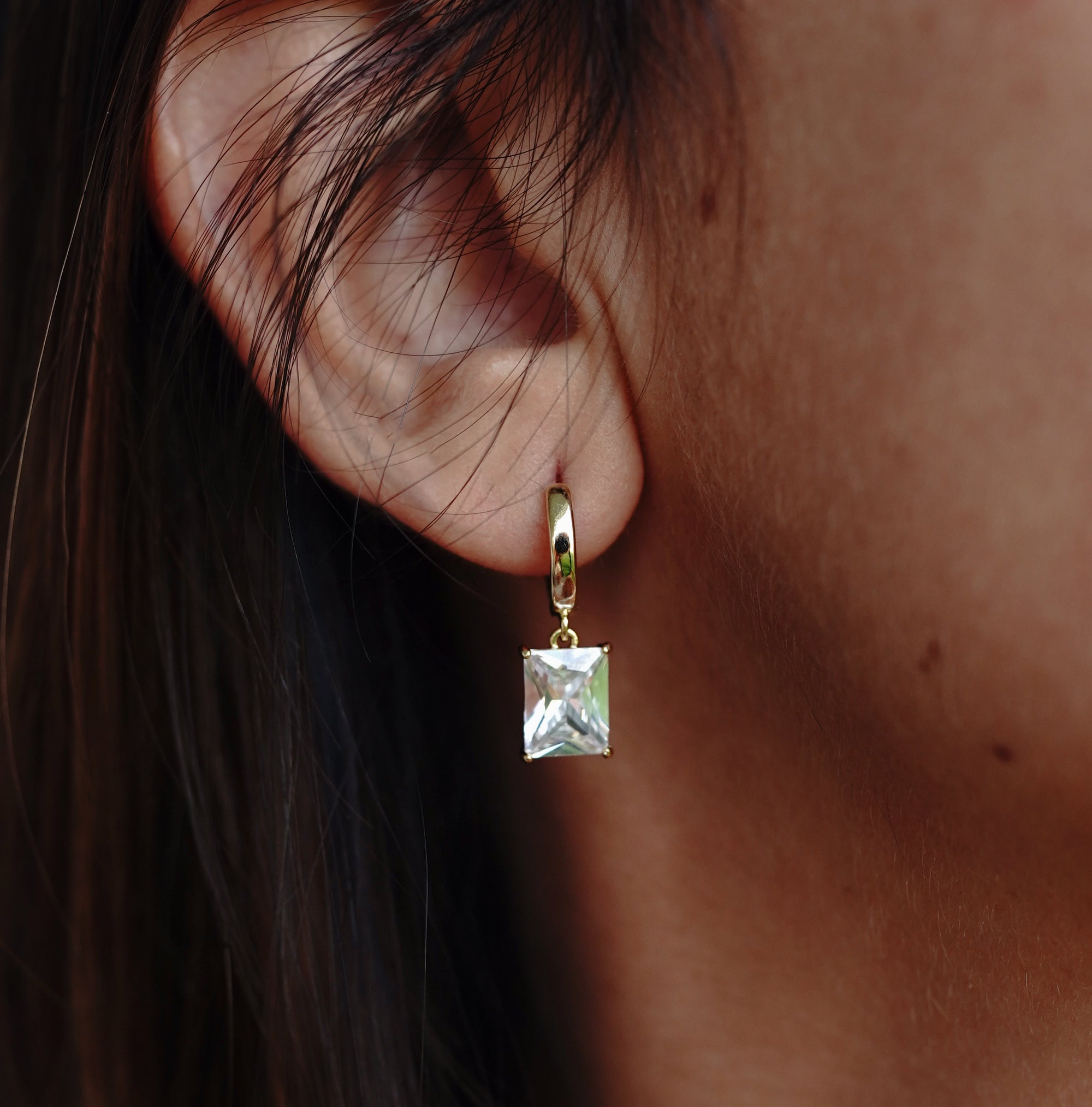 A close-up image of a woman's ear wearing a gold-colored drop earring with a square-shaped zircon stone.