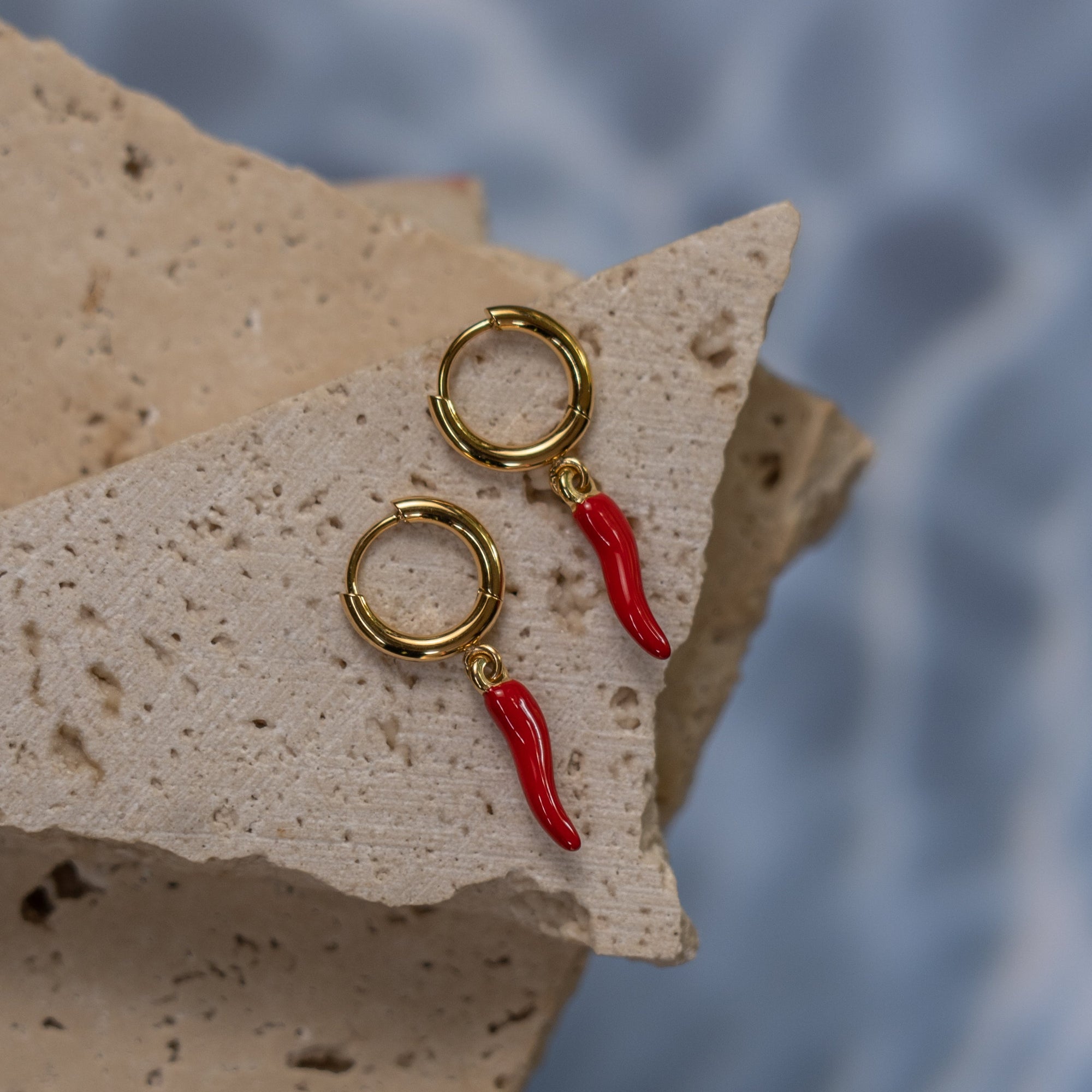 A pair of gold-colored hoop earrings with red chili-shaped drops, displayed on a textured beige surface with a water wave background.
