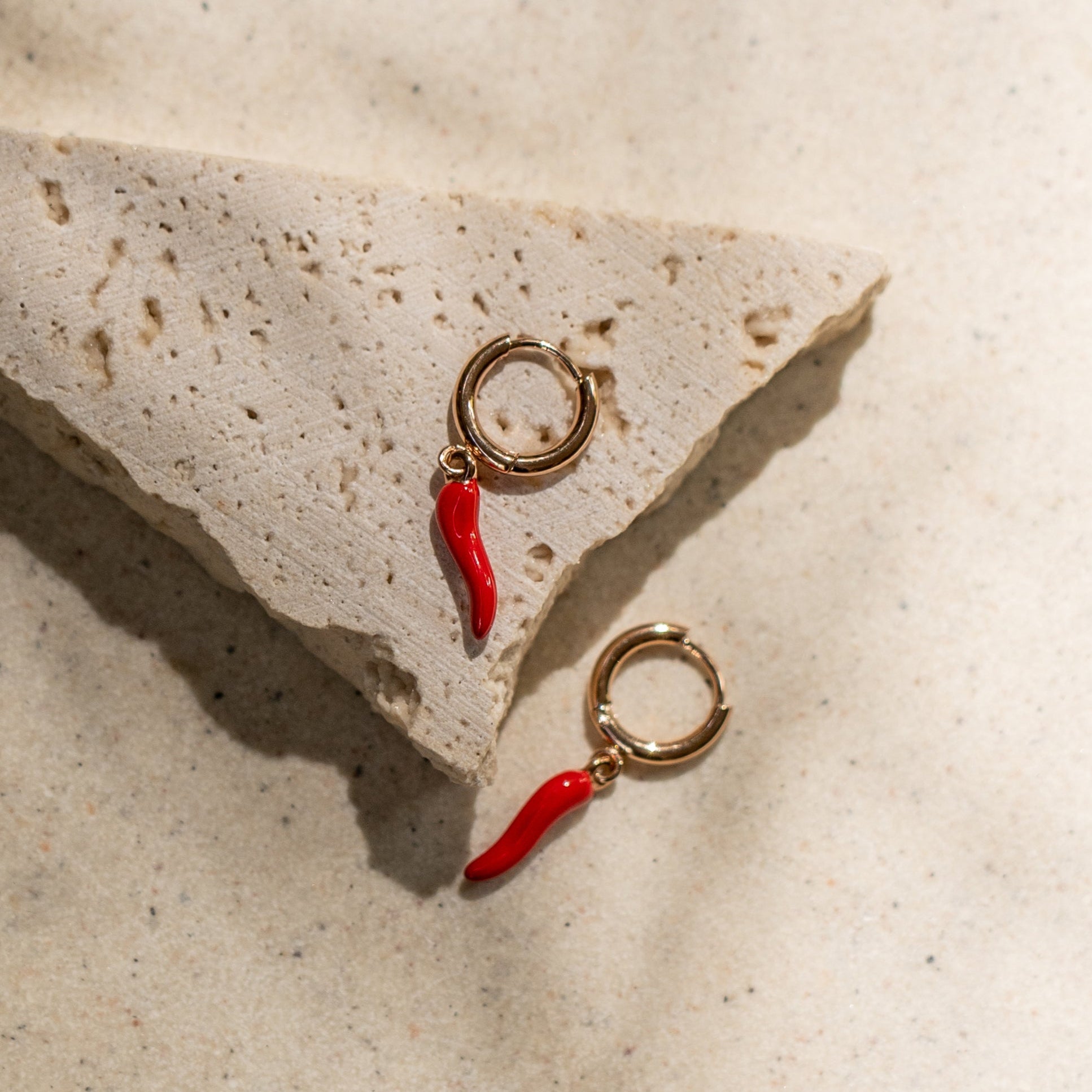 A pair of gold-colored hoop earrings with red chili-shaped drops, displayed on a textured beige surface with a water wave background.