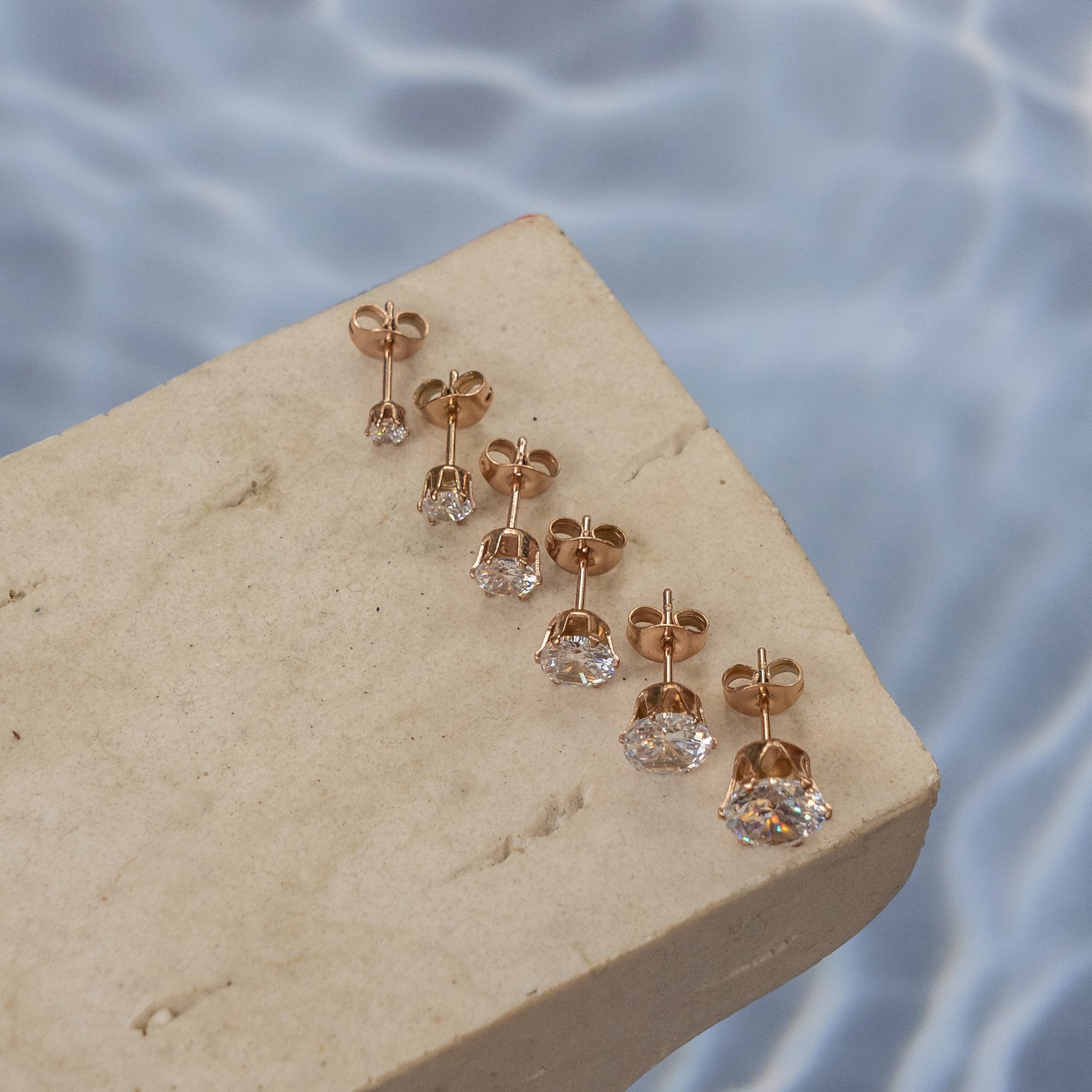 A collection of rose gold Zirconia stud earrings displayed on a light-colored surface with water droplets in the background.