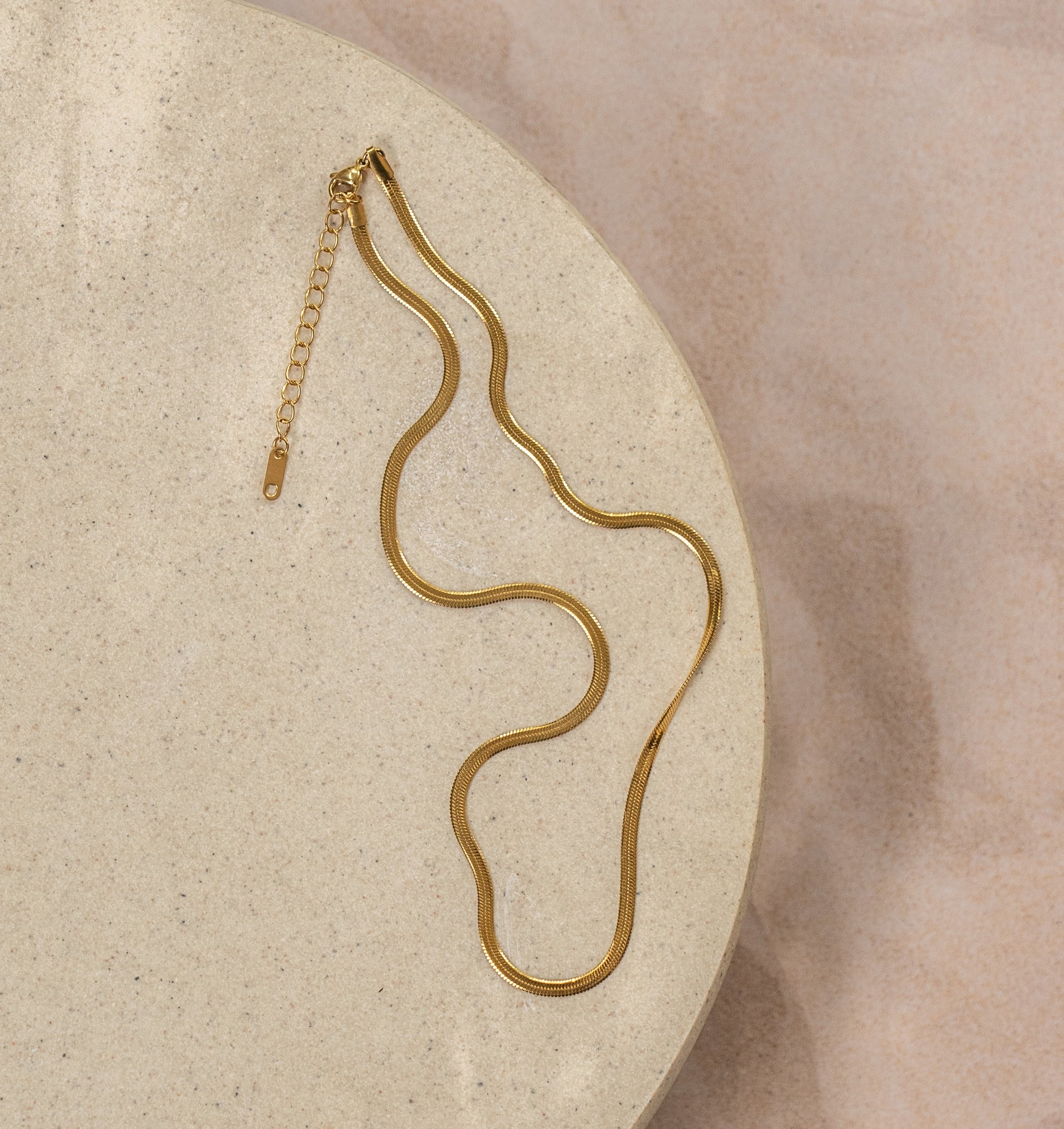 A gold-colored herringbone necklace laid out on a beige surface.