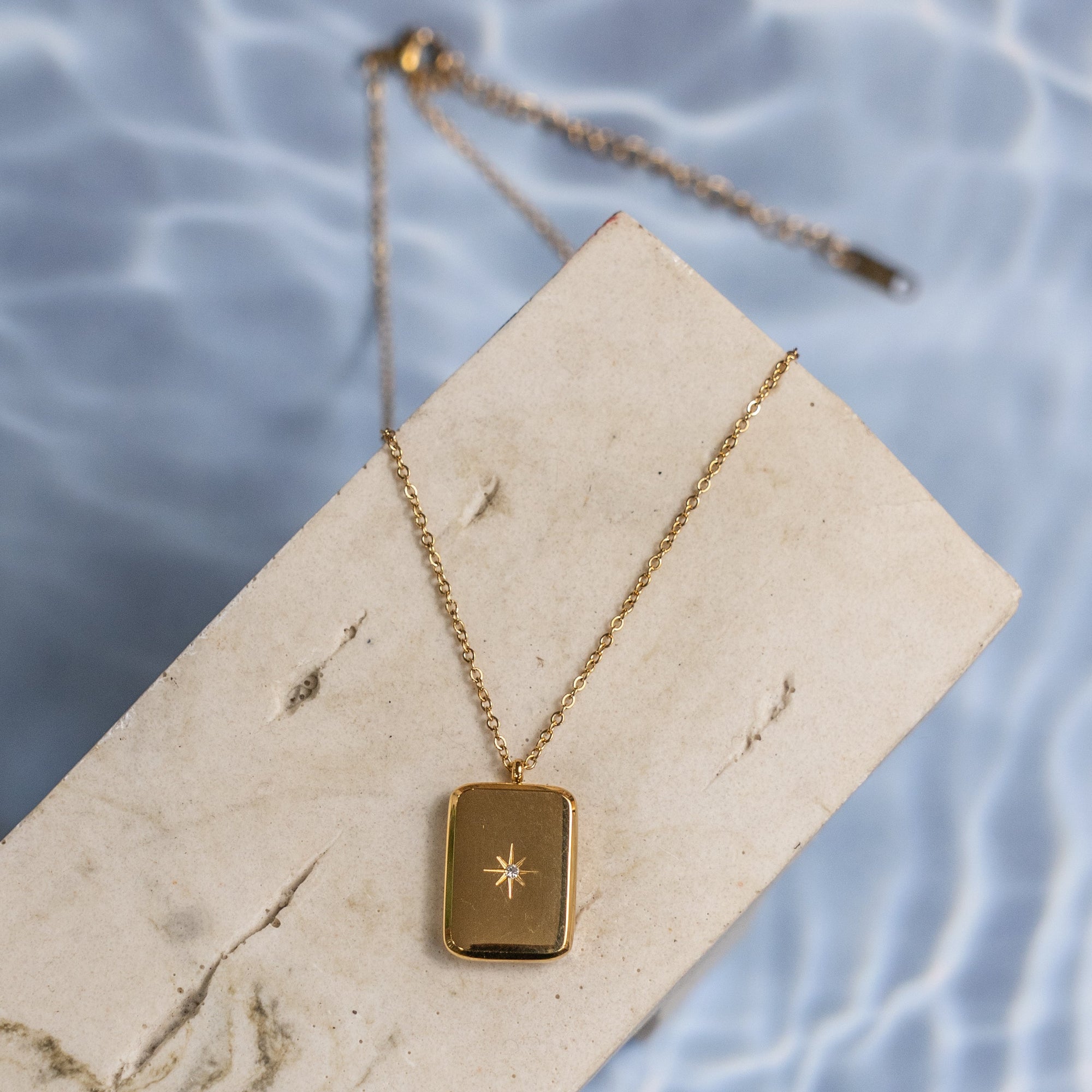 A gold-colored necklace with a small star charm on a rectangular link chain, displayed on a wooden surface with a water reflection.