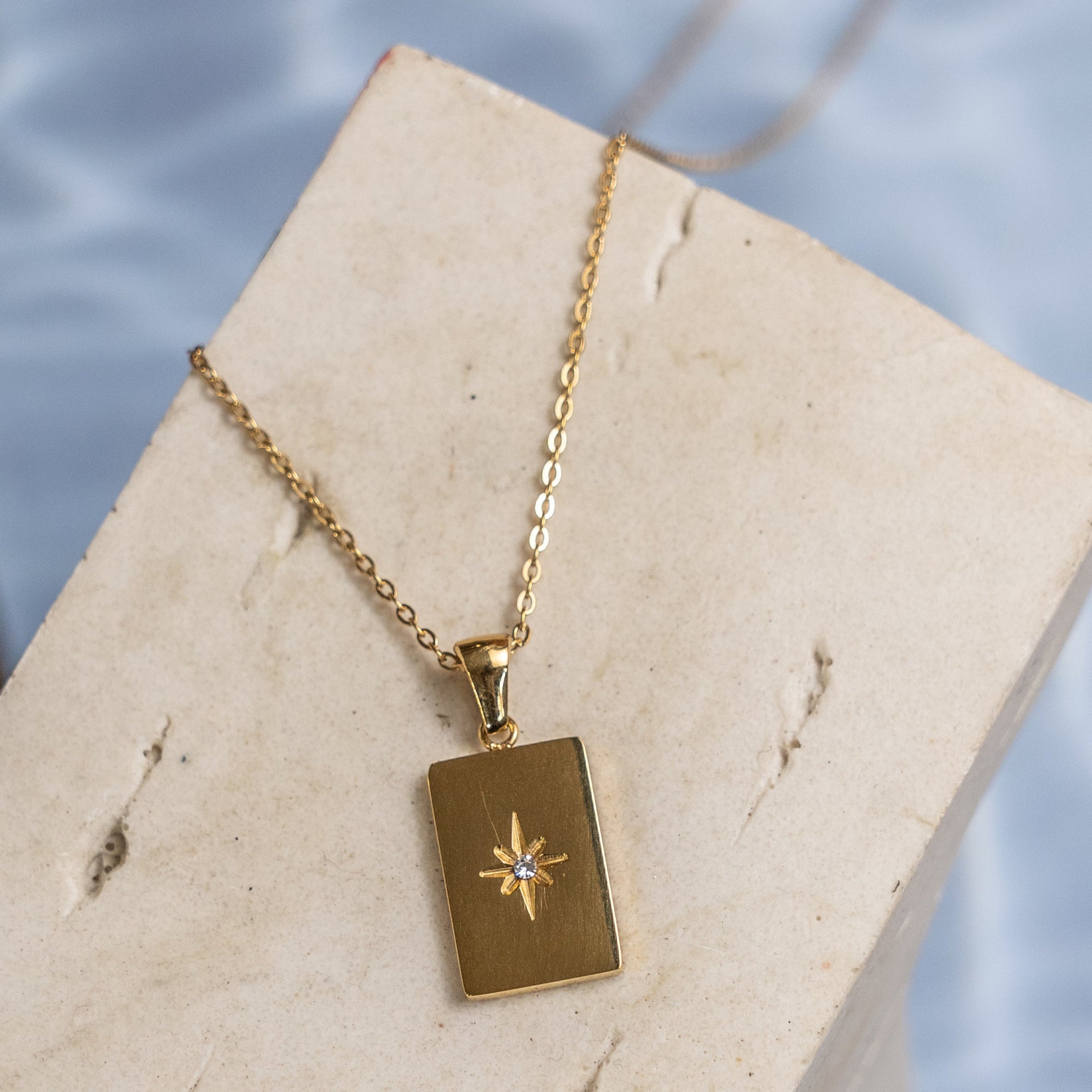 A gold-colored necklace with a rectangular pendant featuring a star design, displayed on a textured white surface.