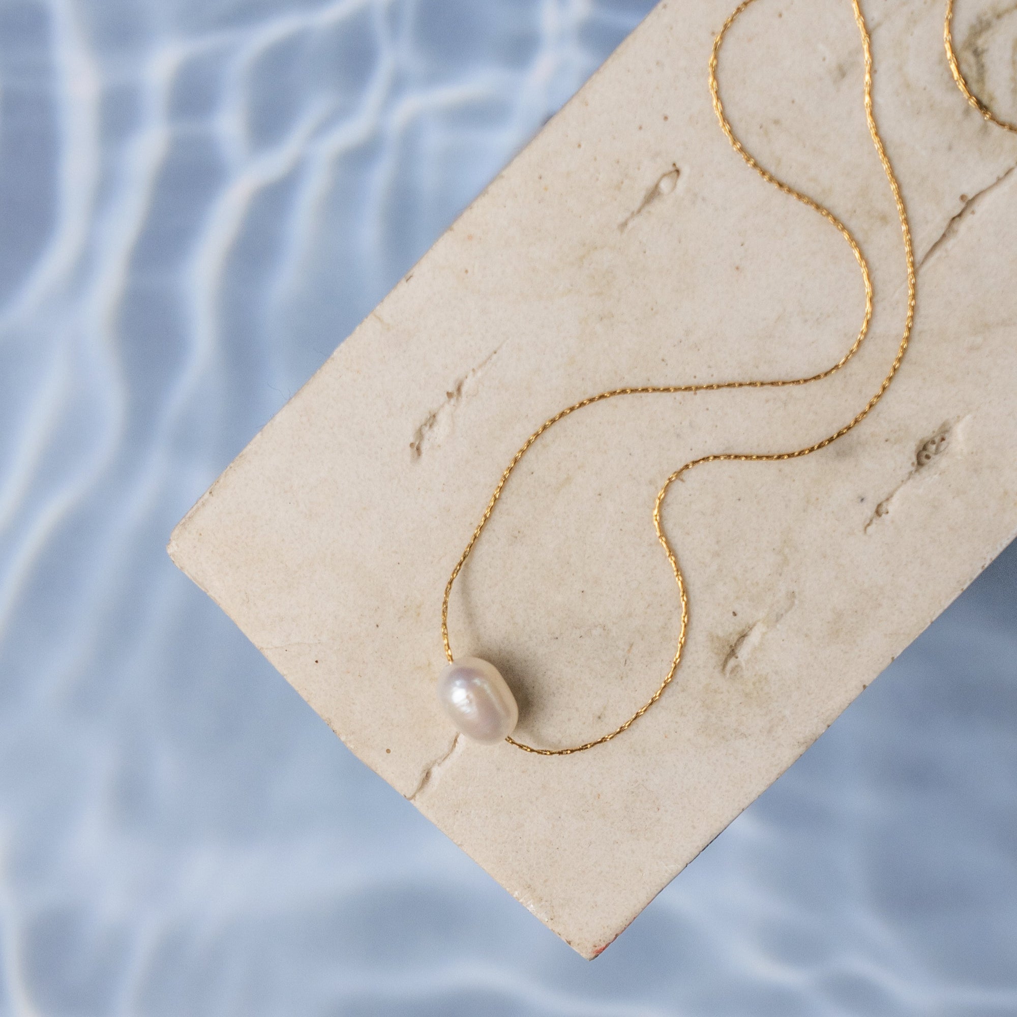 A single freshwater pearl necklace on a gold-tone chain, displayed on a beige surface with water reflection in the background.