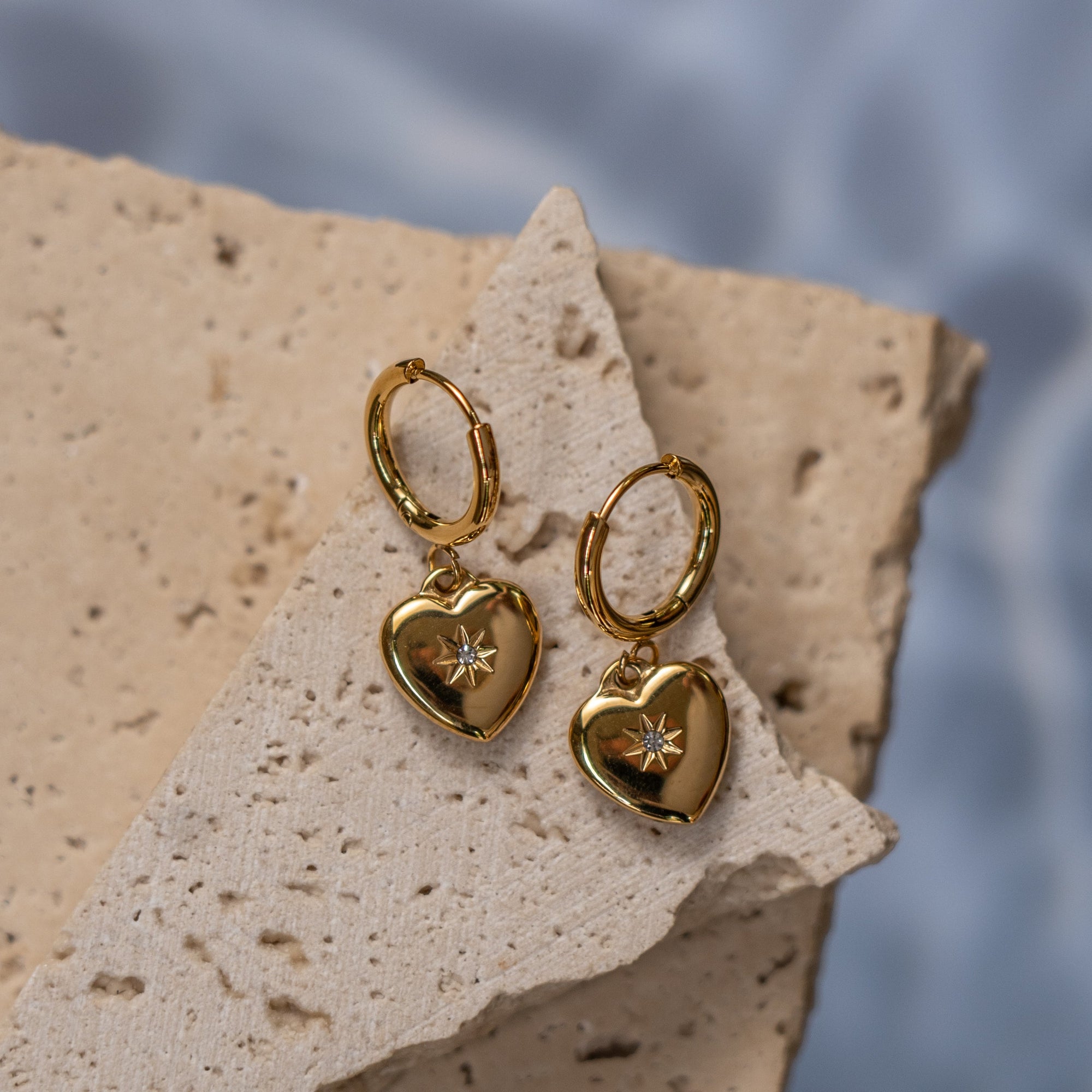 A pair of gold-colored heart-shaped earrings placed on a textured stone surface with water drops in the background.