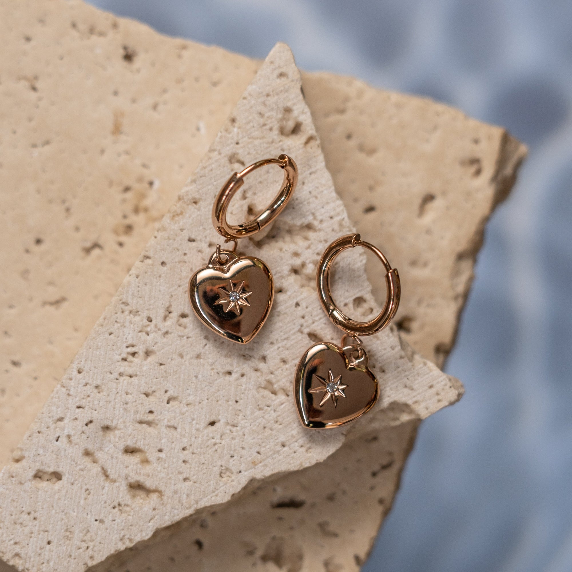 A pair of gold-colored heart-shaped earrings placed on a textured stone surface with water drops in the background.