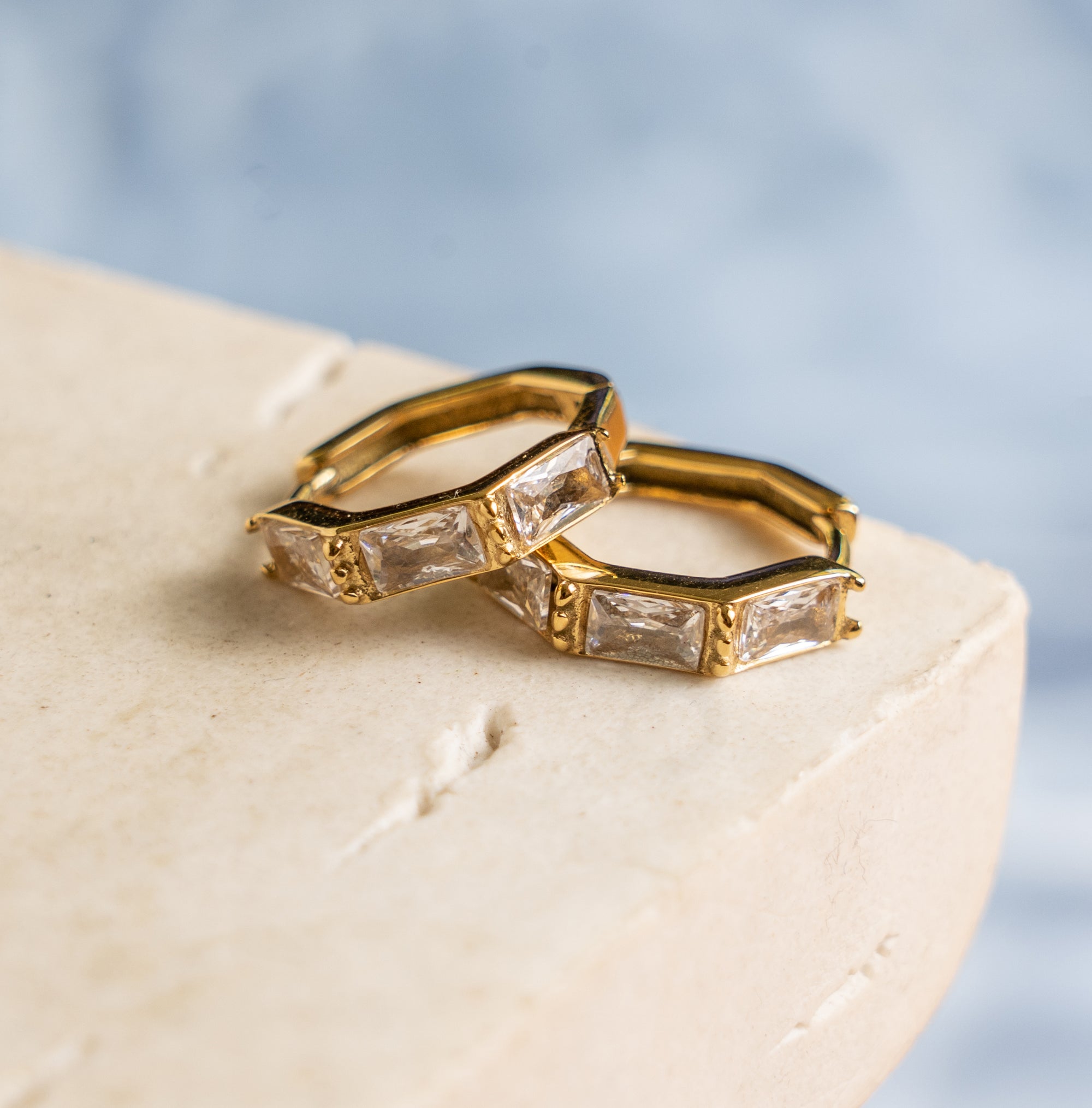 A pair of gold-colored hoop earrings with zirconia stones, displayed on a beige surface.