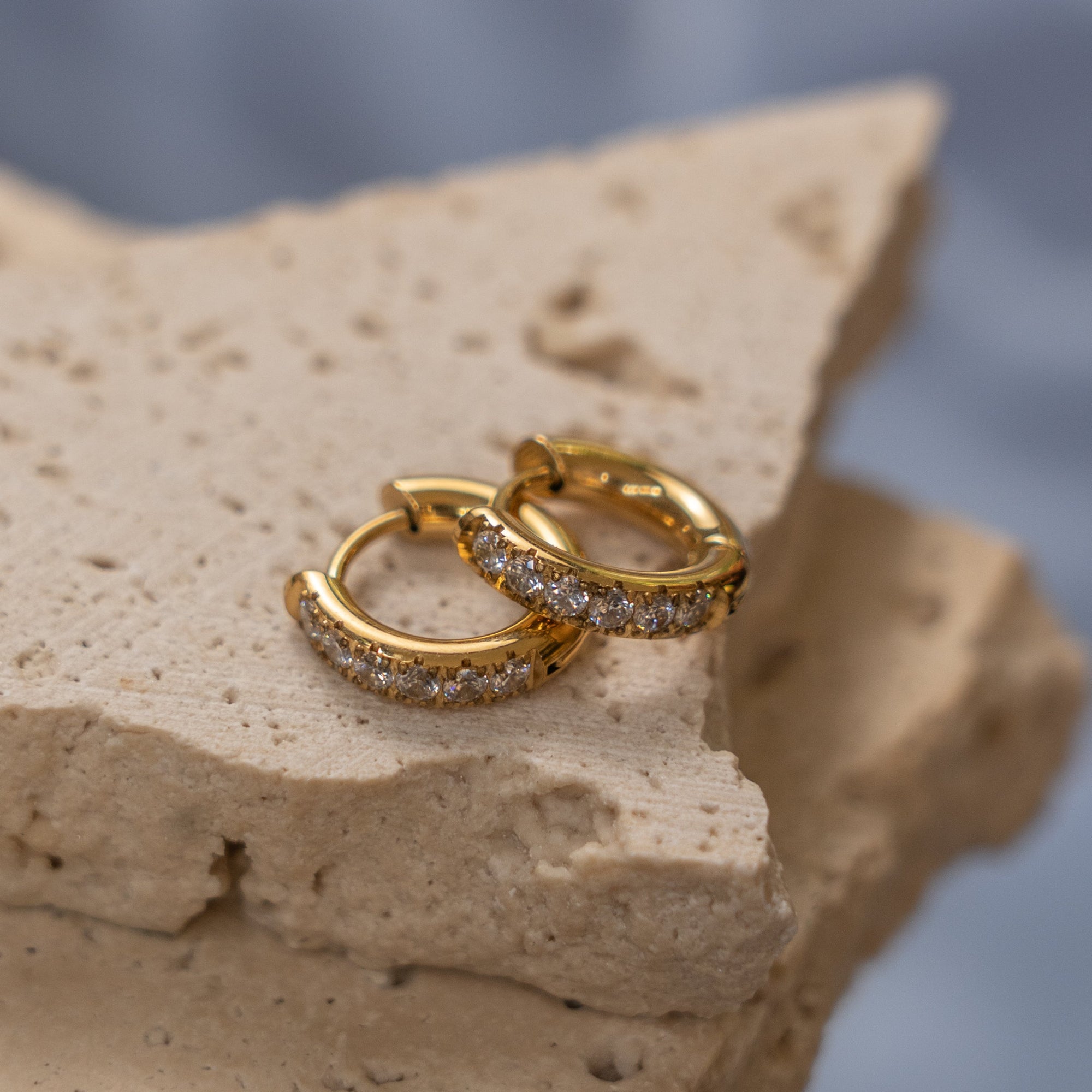 A pair of gold-colored huggie earrings adorned with zirconias, displayed on a textured beige surface.