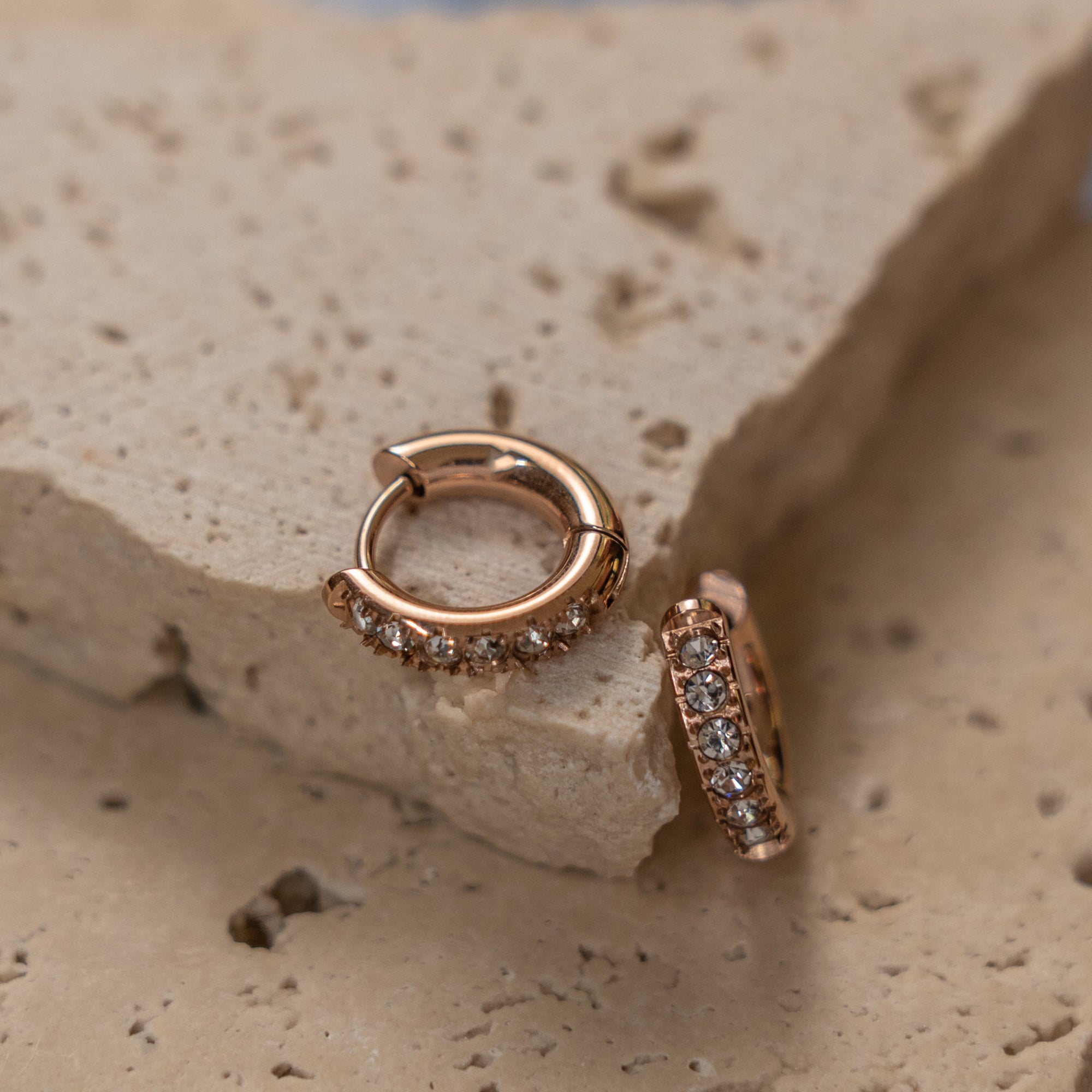 A pair of gold-colored huggie earrings adorned with zirconias, displayed on a textured beige surface.