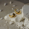 Gold moon-shaped earrings on a textured white surface with pebbles in the background