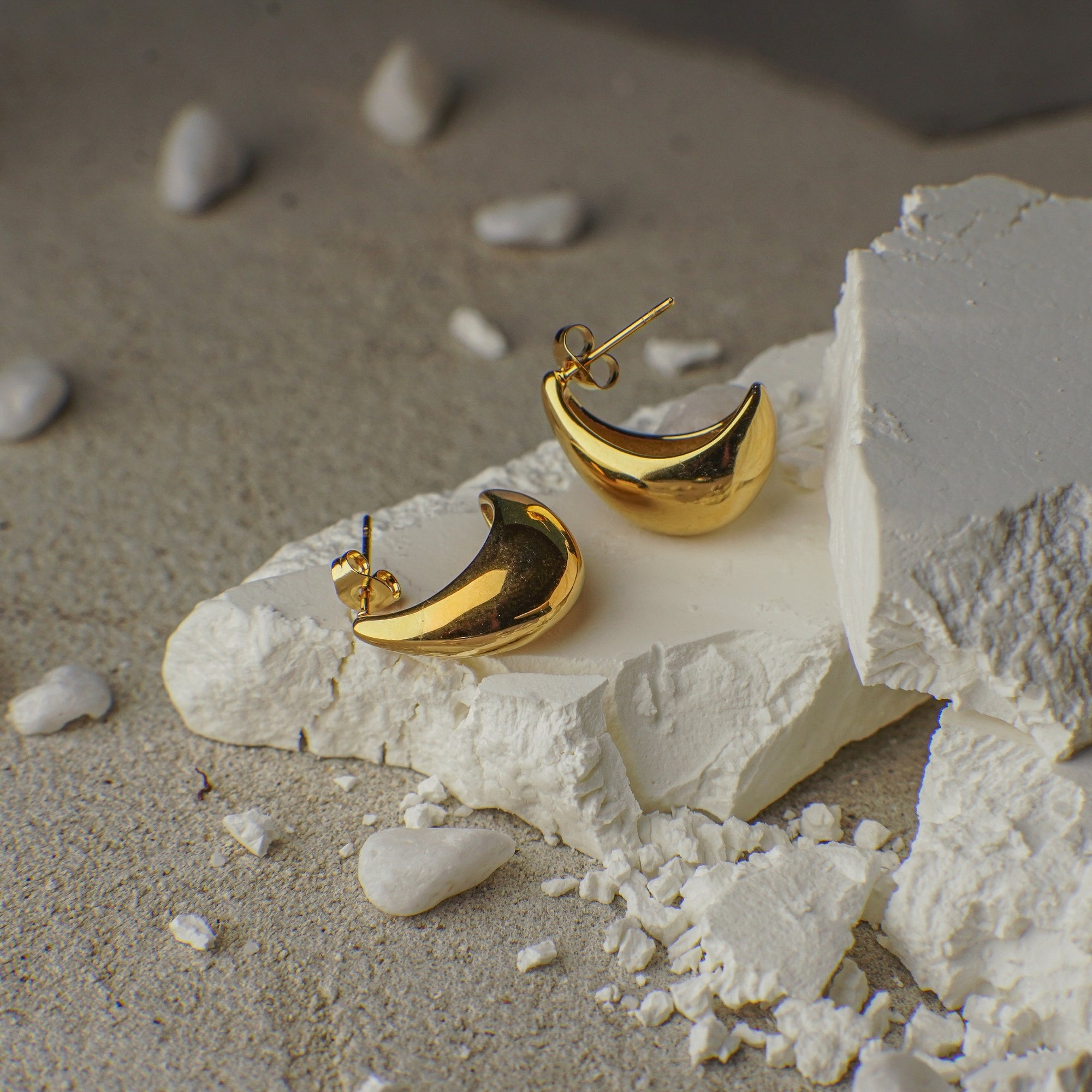 Gold moon-shaped earrings on a textured white surface with pebbles in the background