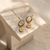 A pair of gold-colored huggie earrings with a small circular charm attached, displayed on a marble surface.