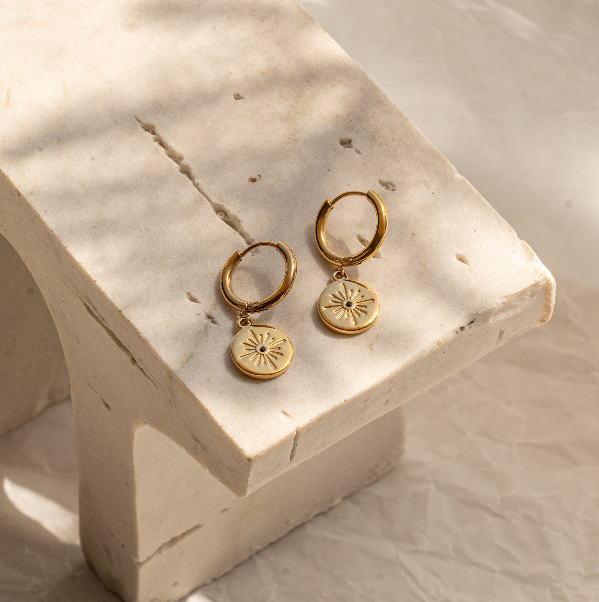 A pair of gold-colored huggie earrings with a small circular charm attached, displayed on a marble surface.