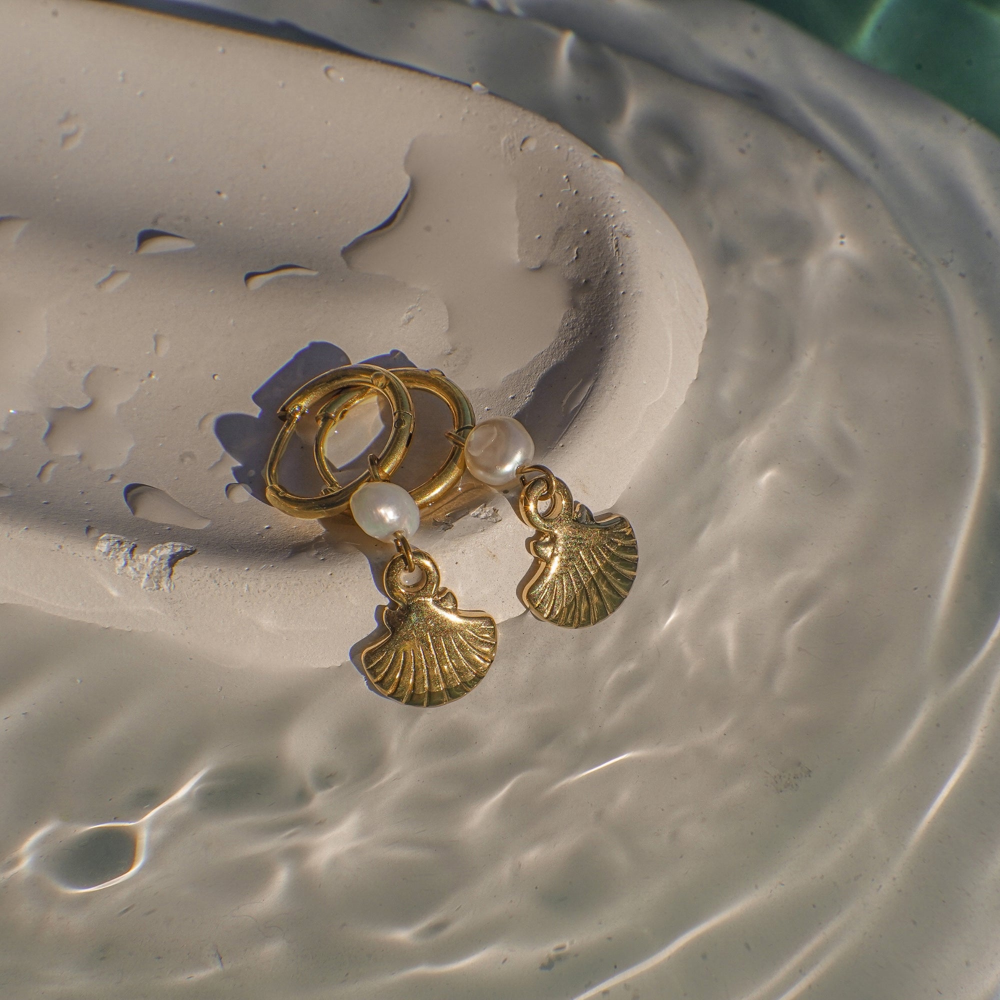 Gold earrings with shell and pearl charms on a textured surface