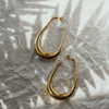 A pair of large gold drop earrings with an abstract design, laid on a crumpled silver paper background.