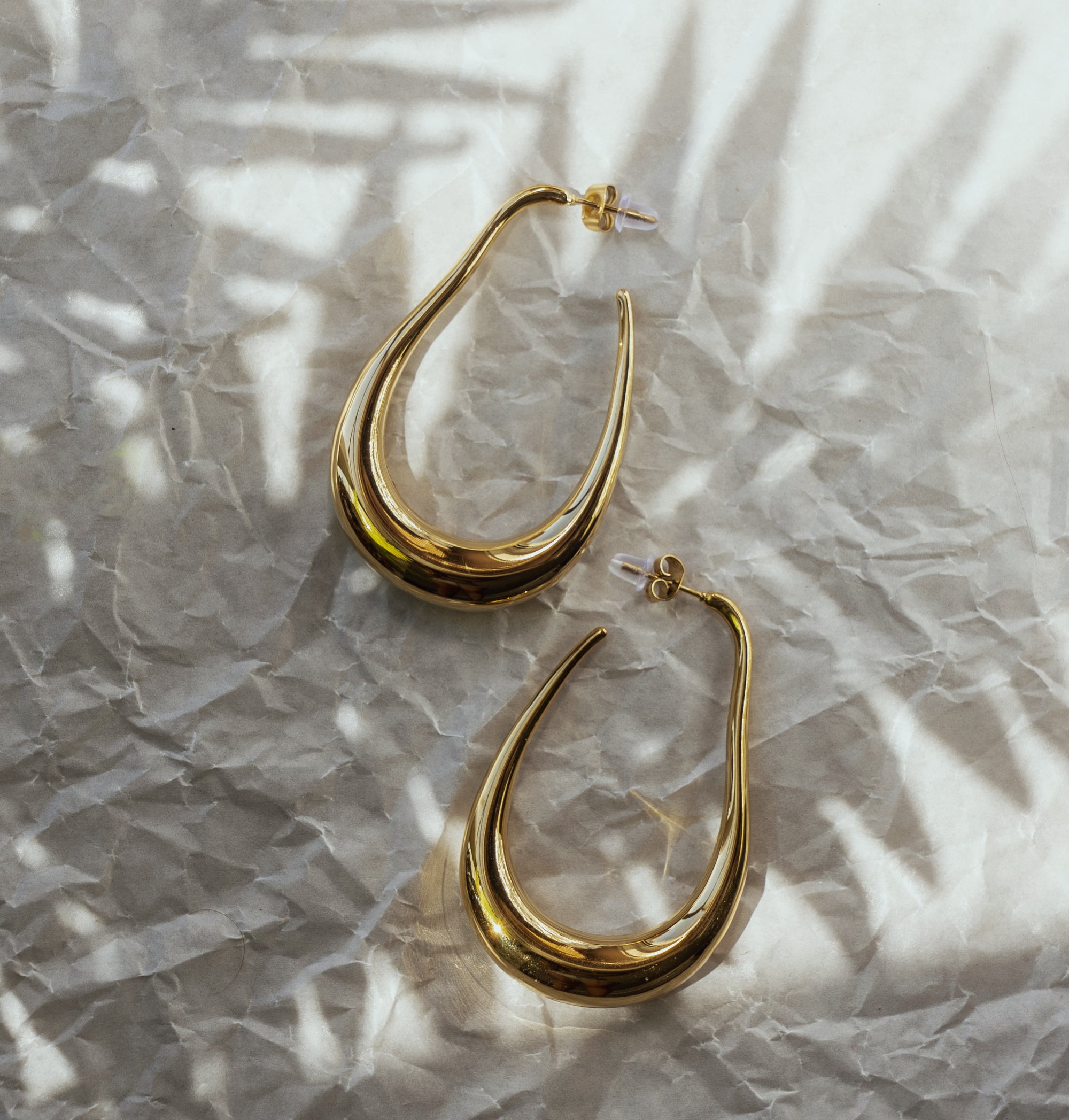 A pair of large gold drop earrings with an abstract design, laid on a crumpled silver paper background.