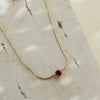 A dainty burgundy necklace with a small, round, red gemstone pendant, displayed on a textured background.