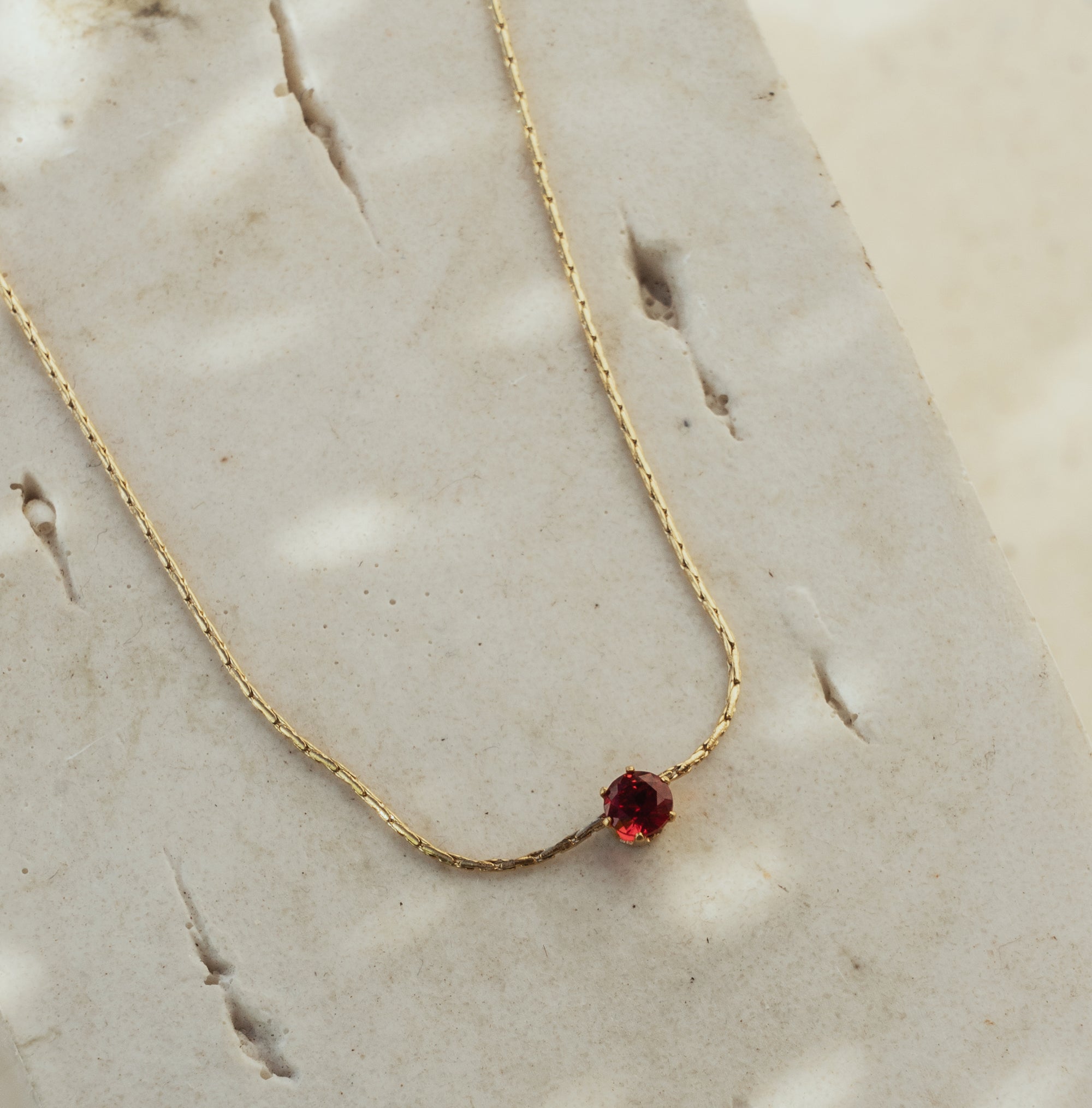 A dainty burgundy necklace with a small, round, red gemstone pendant, displayed on a textured background.
