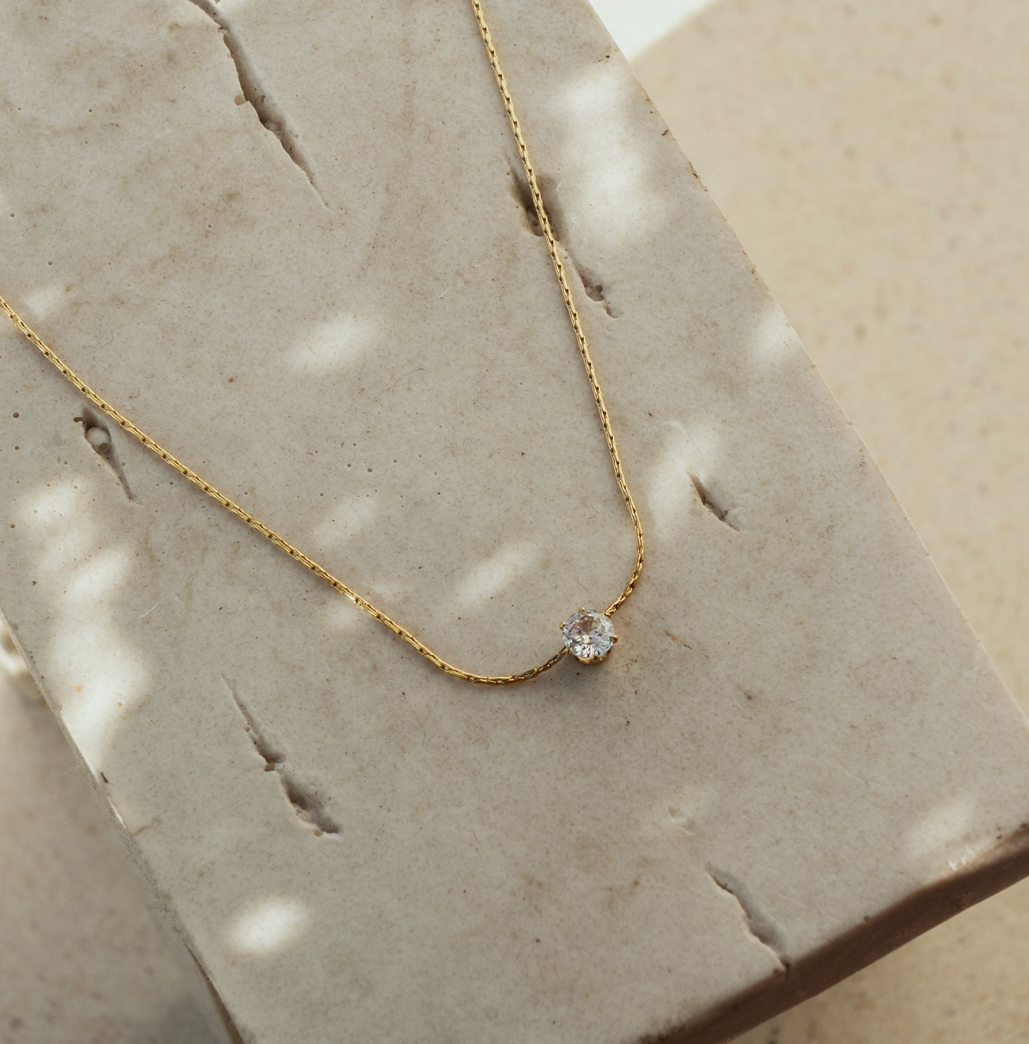 A dainty gold-colored necklace with a single zirconia pendant lying on a textured surface.