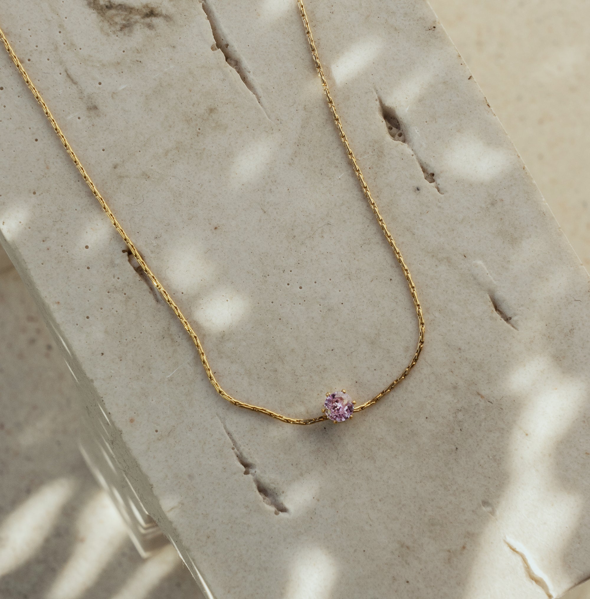 A dainty Aurora Rose Quartz necklace with a gold chain and a pink pendant, laid out on a textured surface.