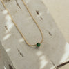 A dainty emerald necklace with a gold chain, displayed on a textured surface.