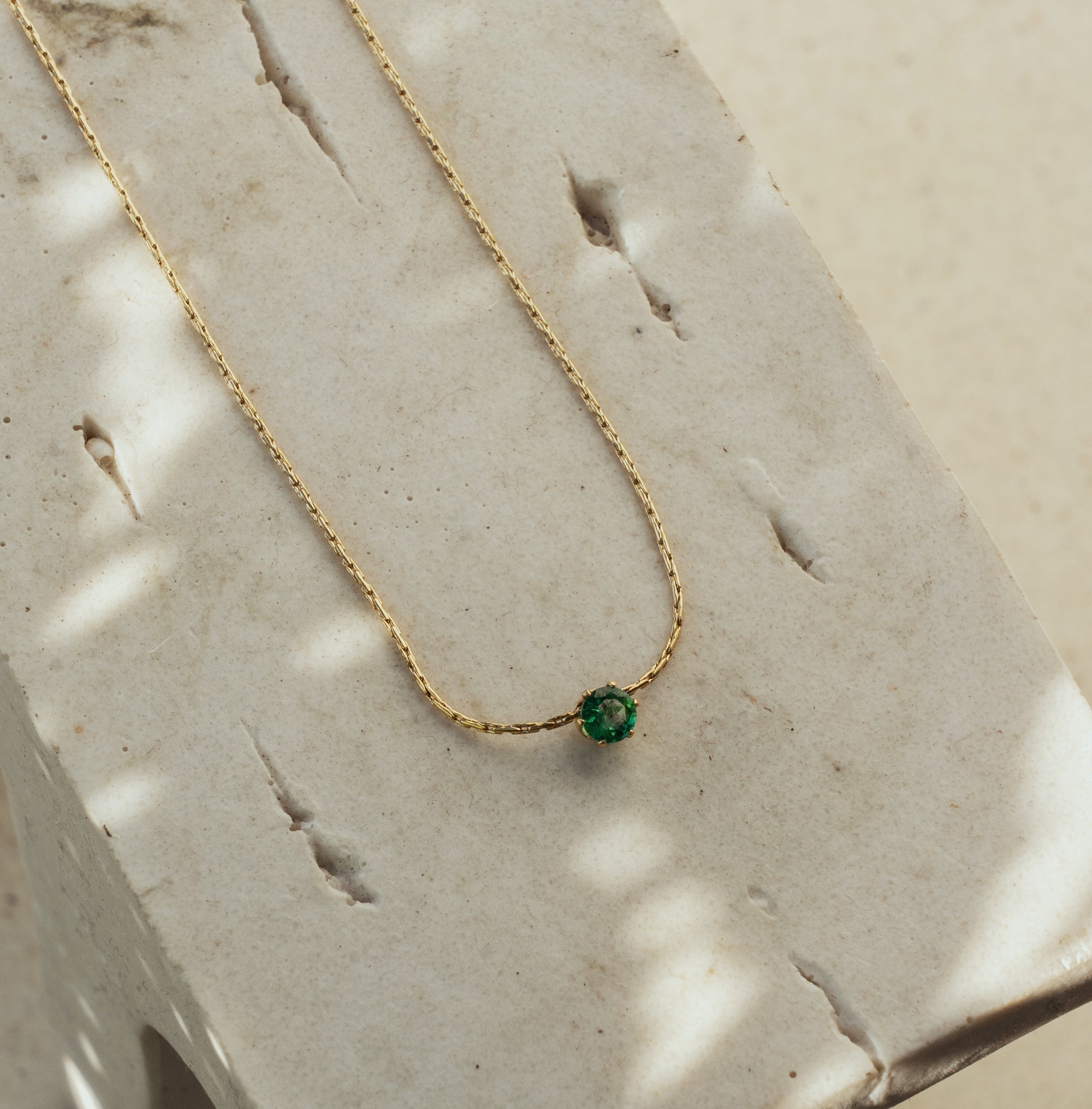 A dainty emerald necklace with a gold chain, displayed on a textured surface.