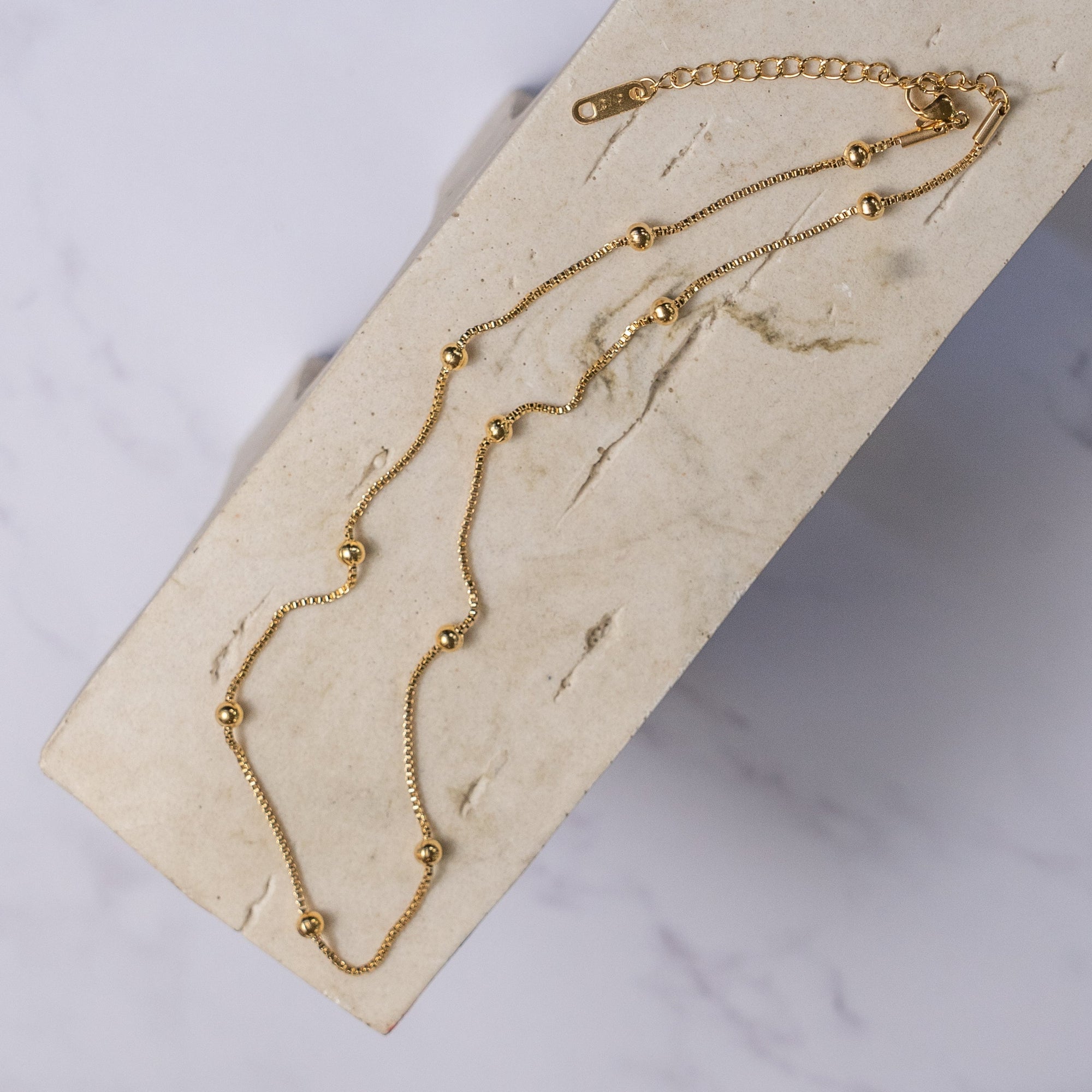 A gold-colored necklace with small regular beads placed along its length, laid out on a beige surface.