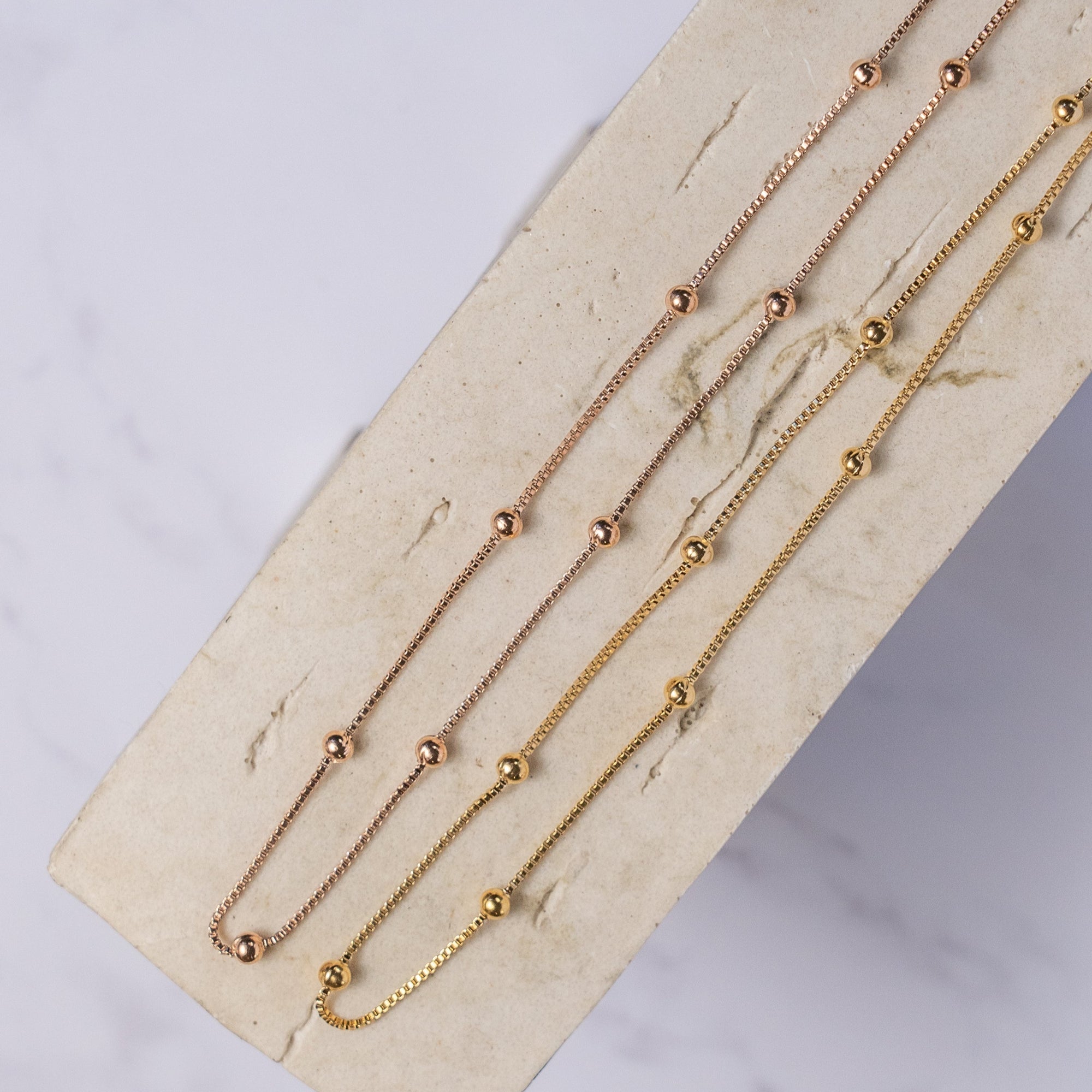 Rose gold and gold-colored necklaces with small regular beads placed along its length, laid out on a beige surface.