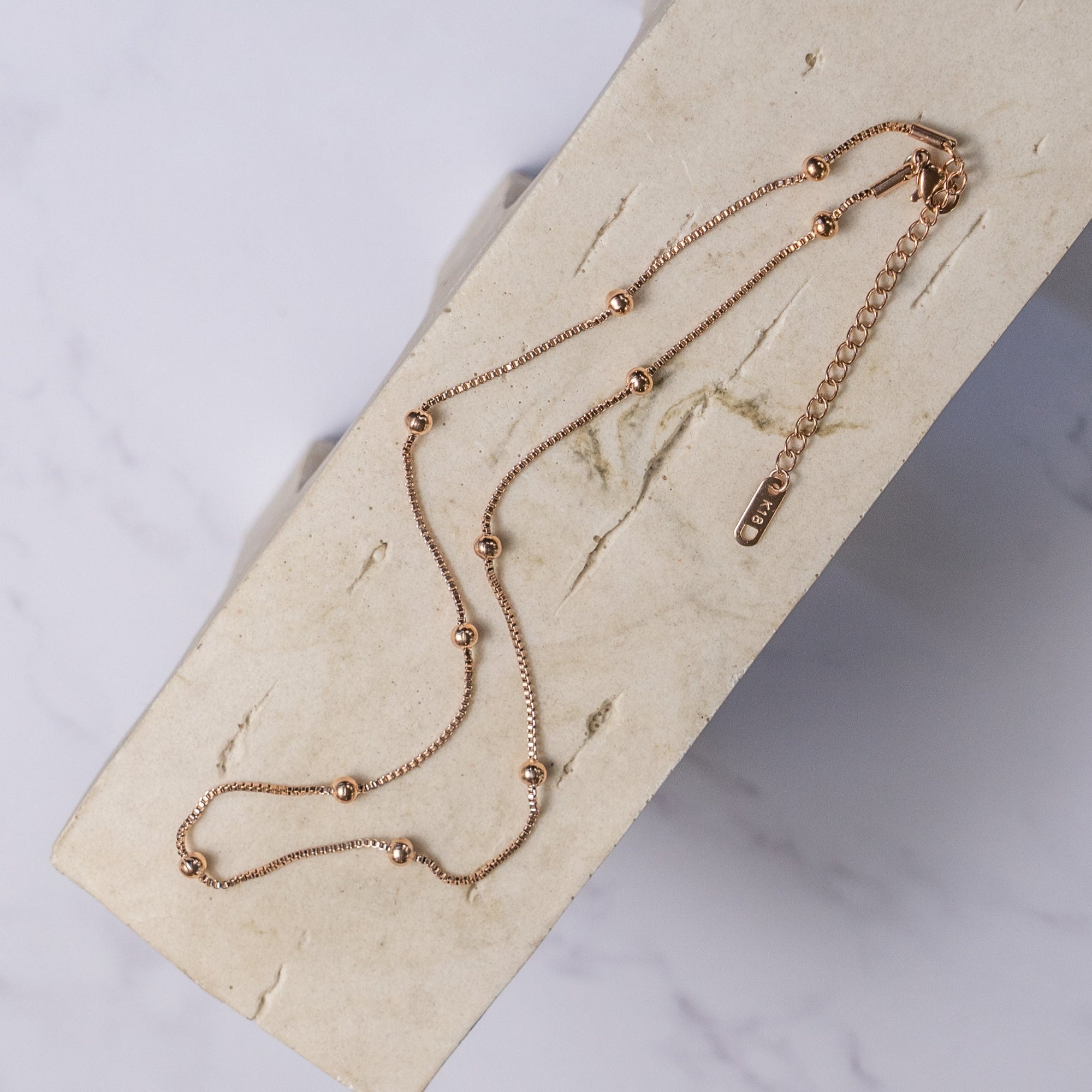 A rose gold-colored necklace with small regular beads placed along its length, laid out on a beige surface.