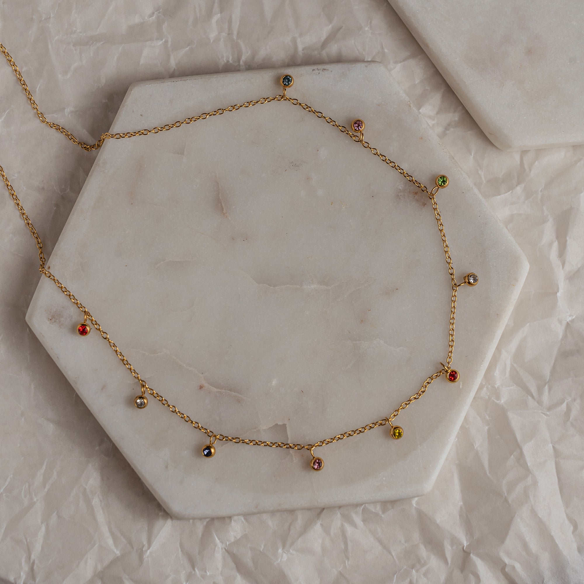 A geometric-patterned necklace with gold-colored metal chain and multicolored zirconia stones laid out on a marble surface.