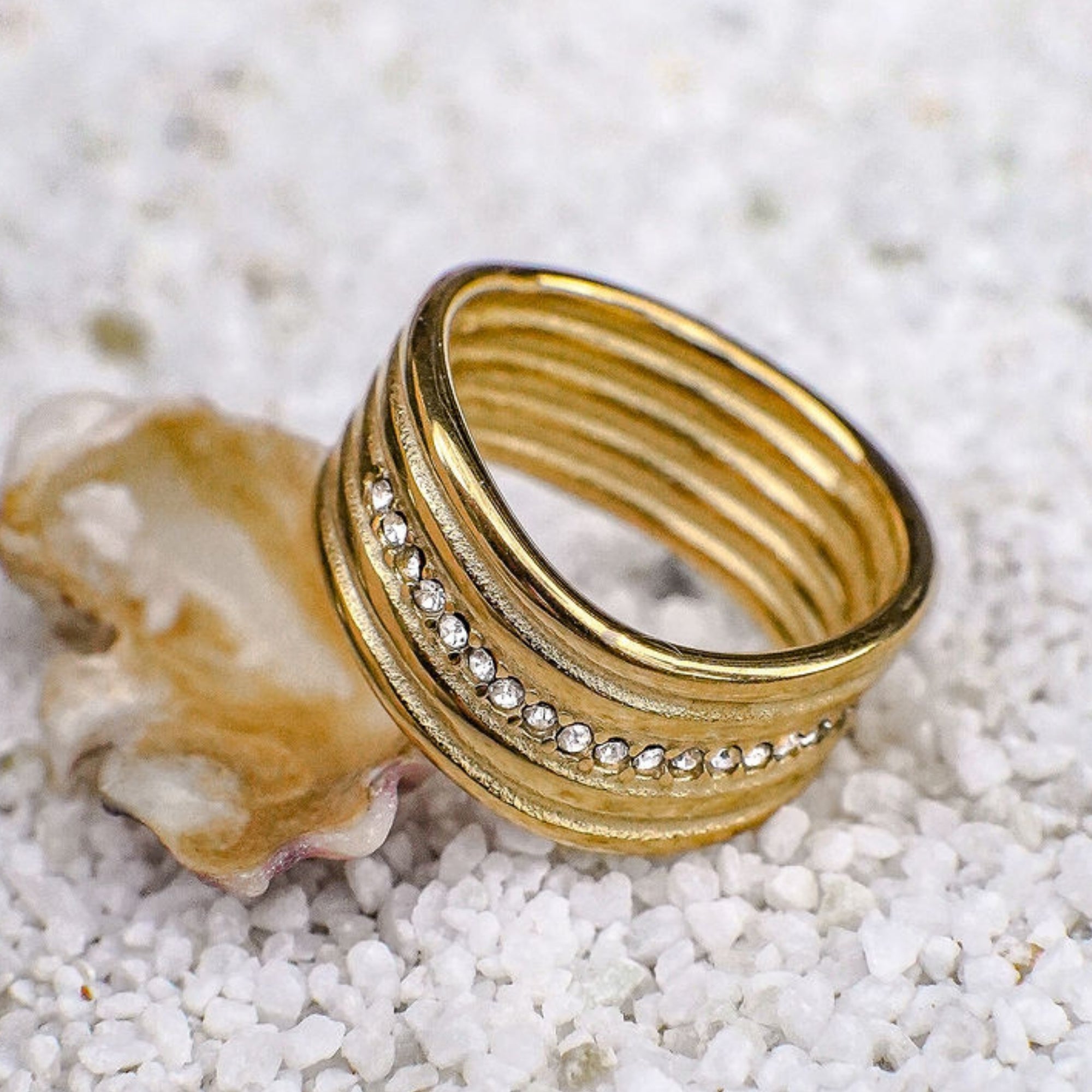 Gold ring with textured design and small stones on a white pebble surface