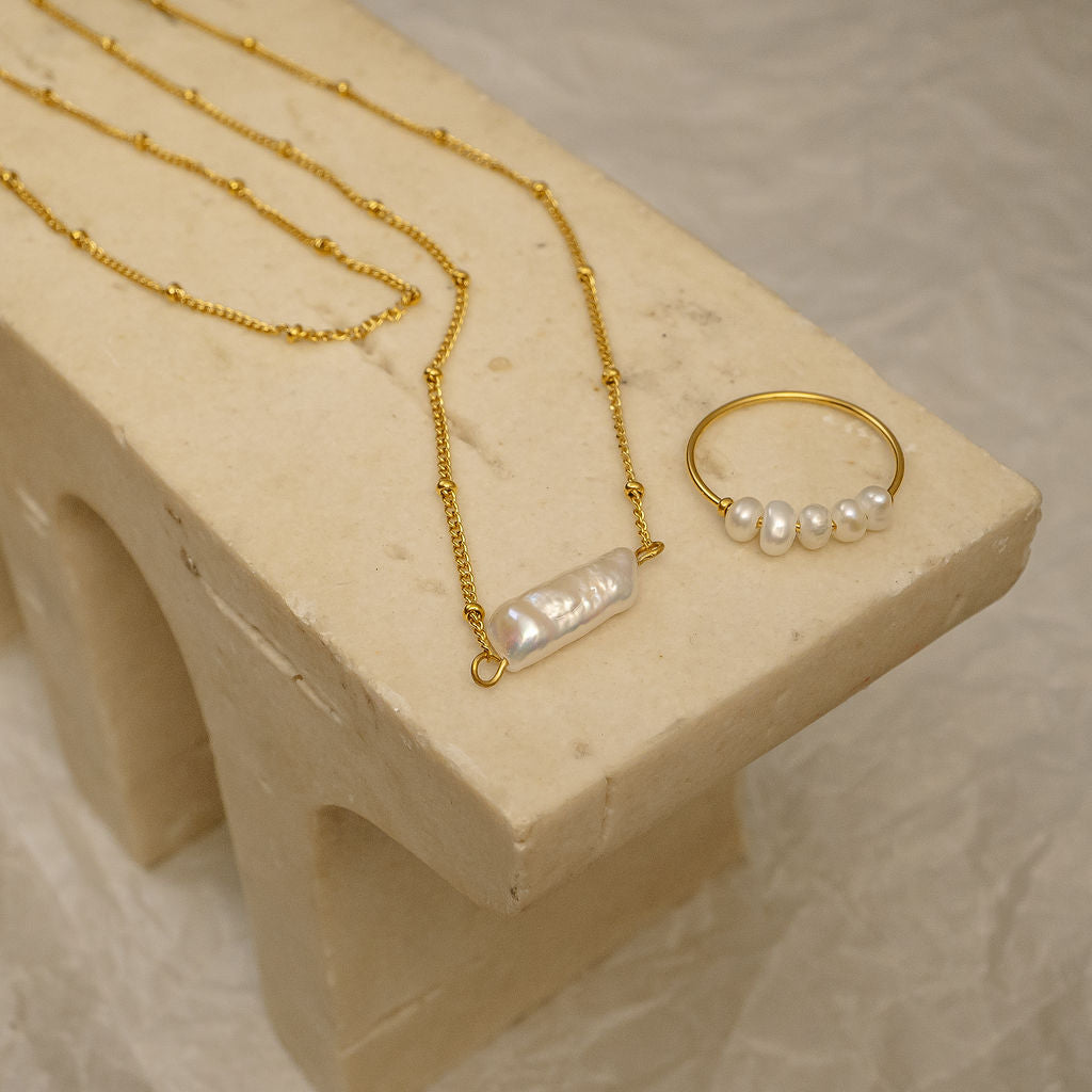 Gold necklace and ring with pearl accents on a beige stone surface