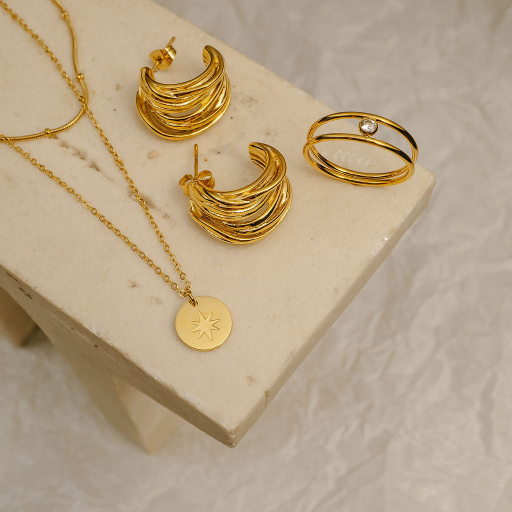 Gold jewelry set including necklace, hoop earrings, and ring on a beige surface.