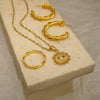 Gold jewelry set including necklace, ring, and hoop earrings on a beige stone surface.