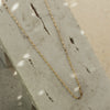 A gold-colored necklace laid out on a textured surface, likely representing a loop-de-loop chain design.