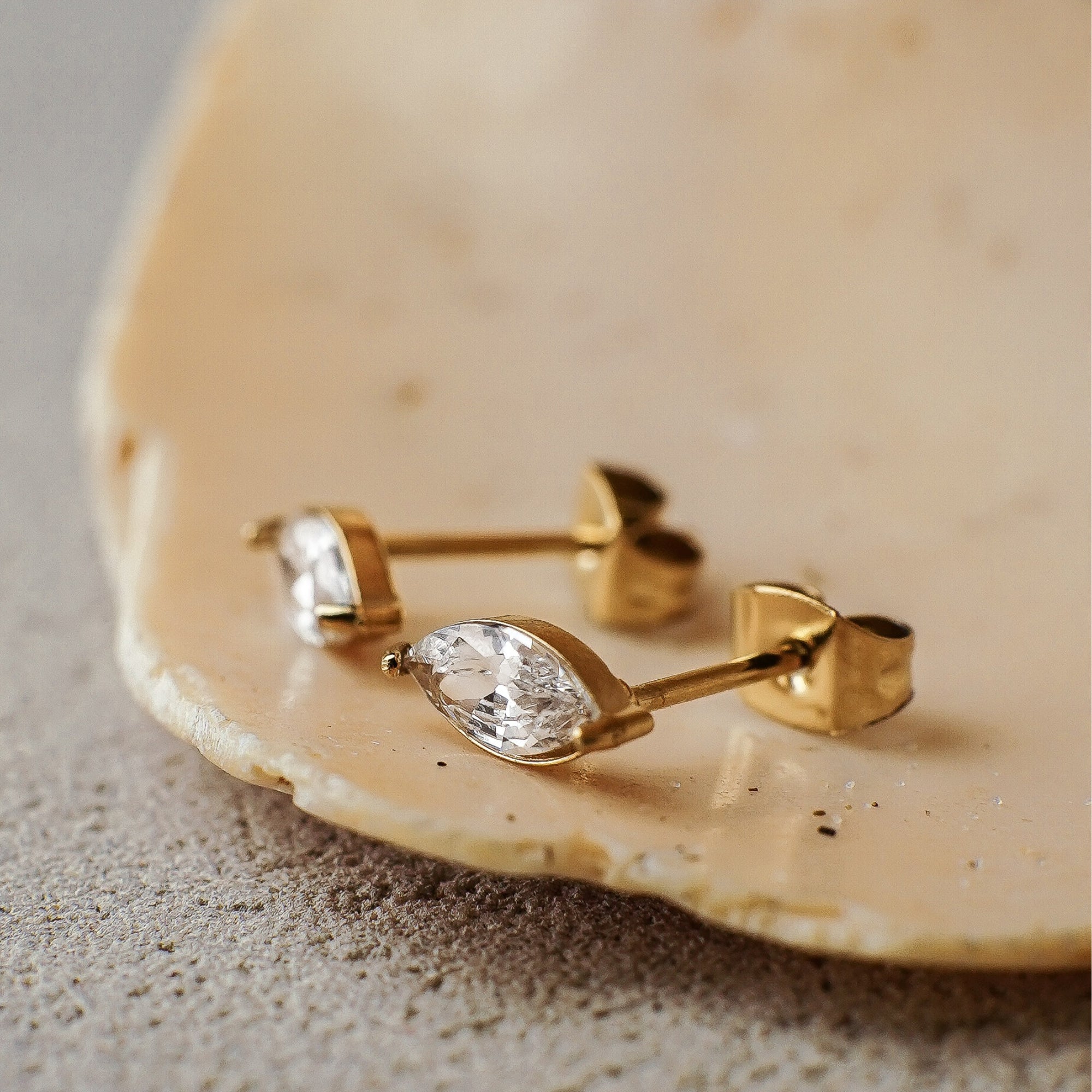 Gold earrings with clear gemstones on a beige stone surface