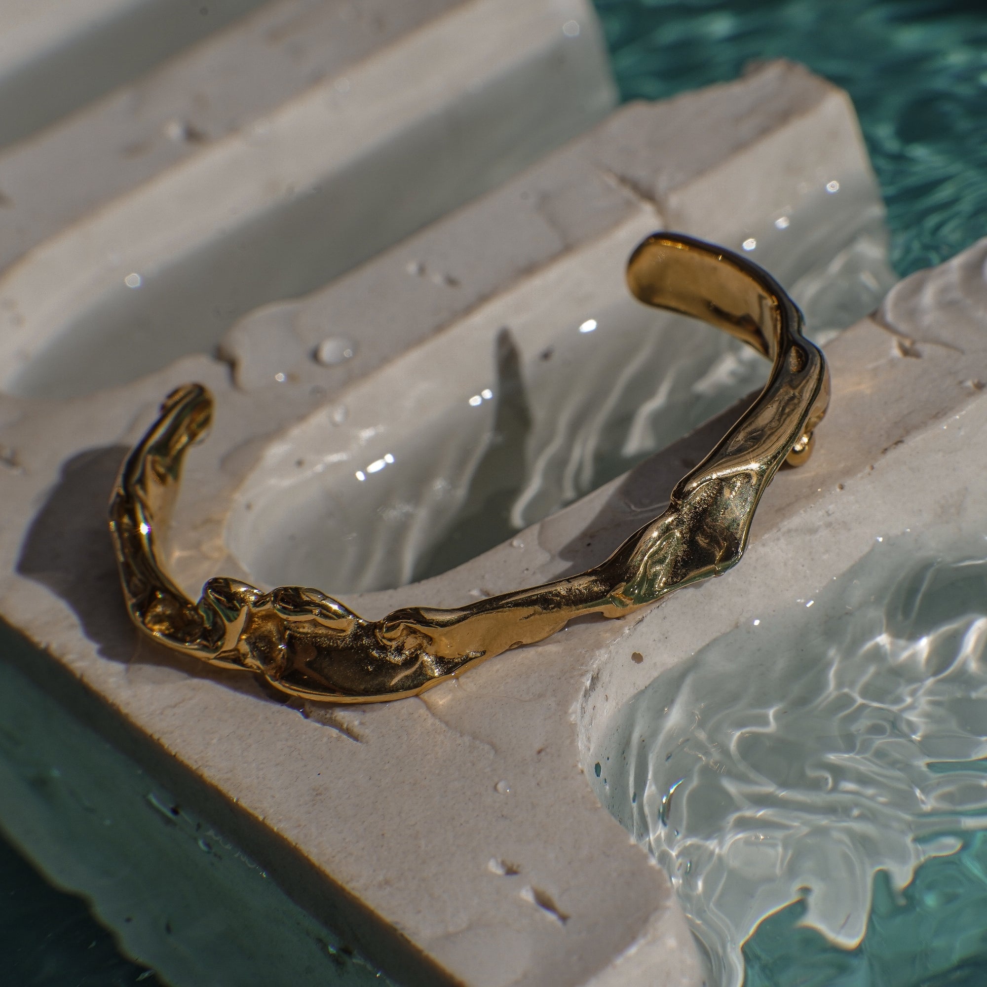Curvy gold bracelet on a stone ledge with water in the background