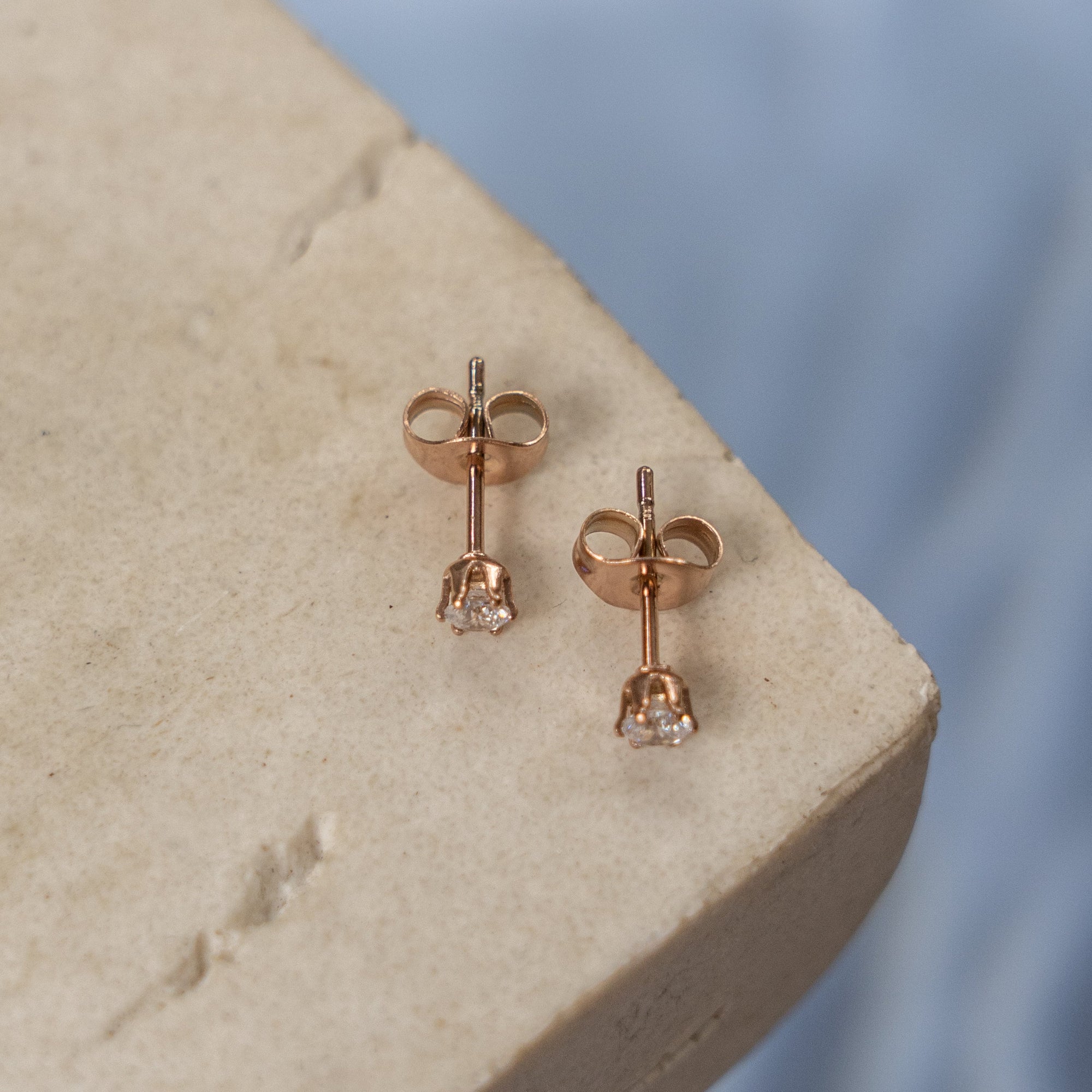 A collection of rose gold Zirconia stud earrings displayed on a light-colored surface with water droplets in the background.