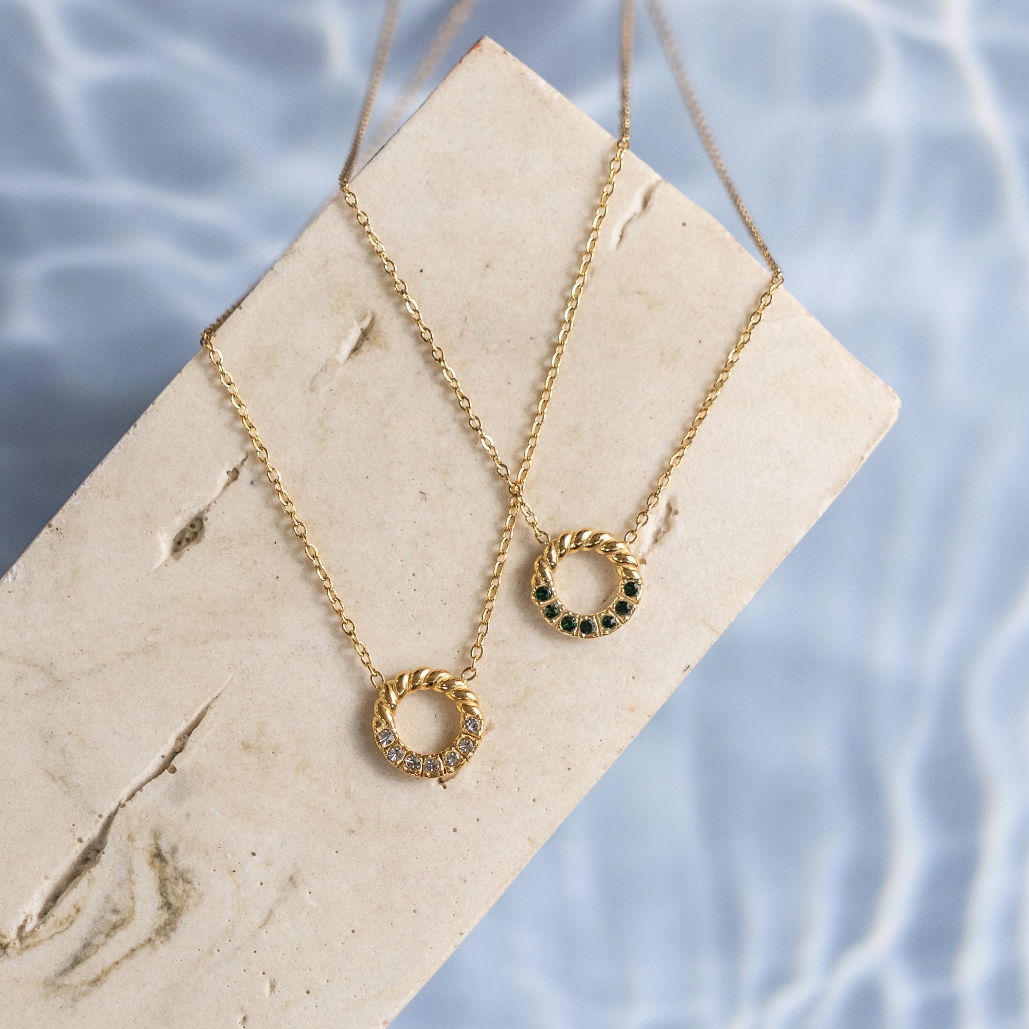 Two gold necklaces with circular pendants on a stone surface with water in the background