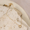 A gold-colored necklace with a small circular zirconia pendant, displayed on a beige rock against a beige textured background.