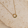 A gold-colored necklace with an emerald green zirconia pendant, displayed on a textured beige surface.