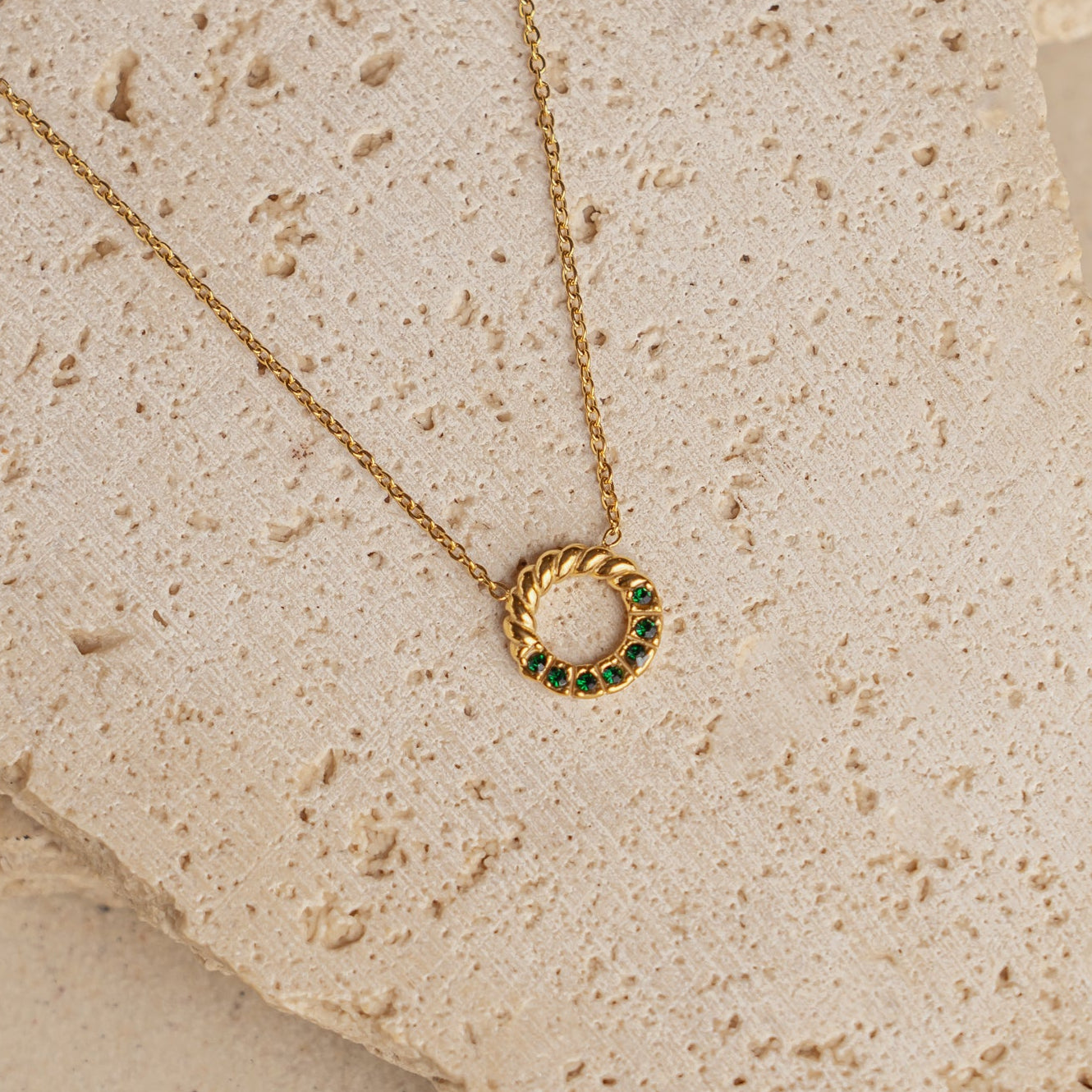 A gold-colored necklace with an emerald green zirconia pendant, displayed on a textured beige surface.