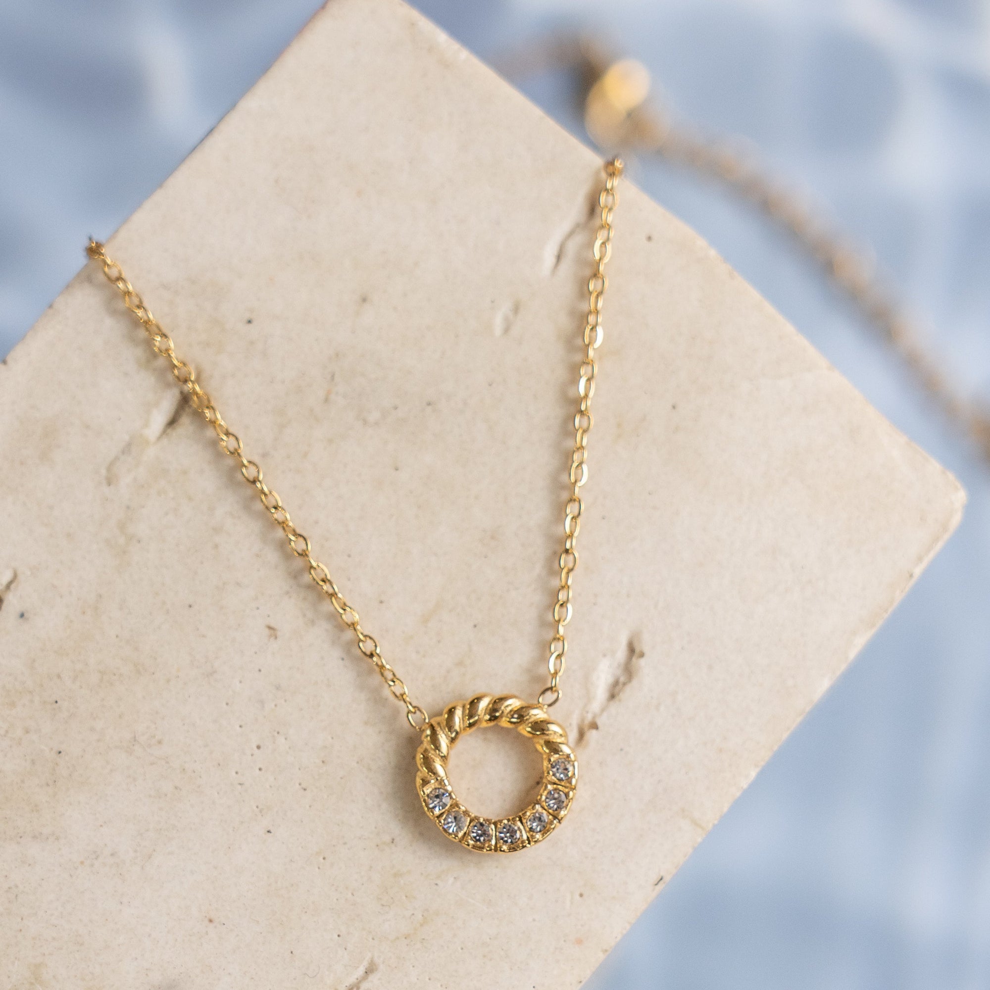 A gold-colored necklace with a small circular zirconia pendant, displayed on a beige card against a blue background.