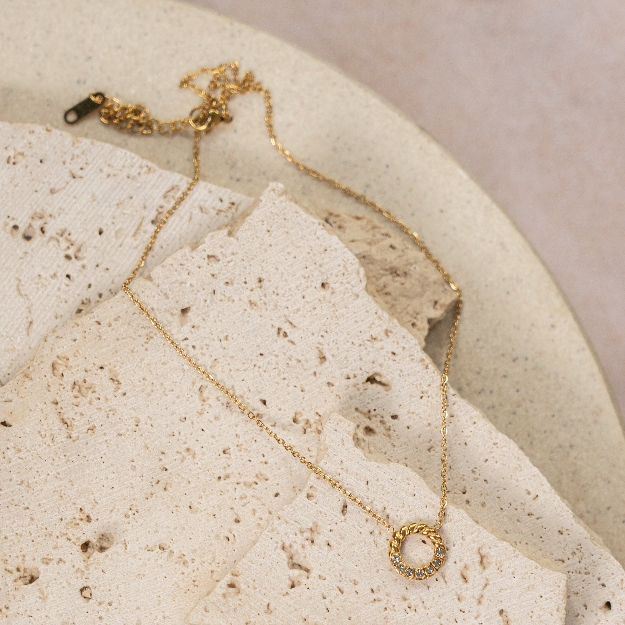 A gold-colored necklace with a small circular zirconia pendant, displayed on a beige rock against a beige textured background.