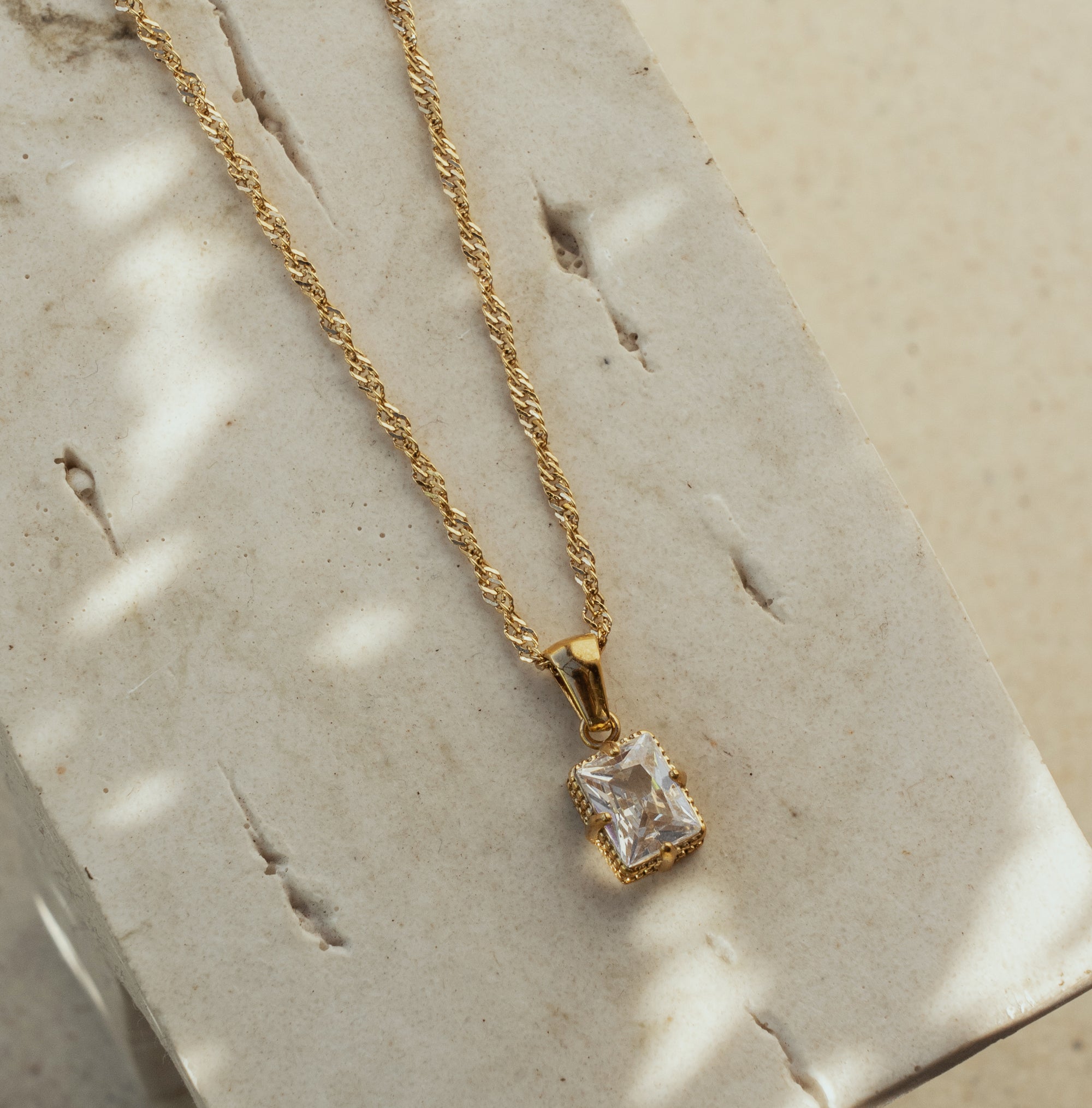 A rose quartz zirconia pendant necklace with a golden chain displayed on a textured surface.