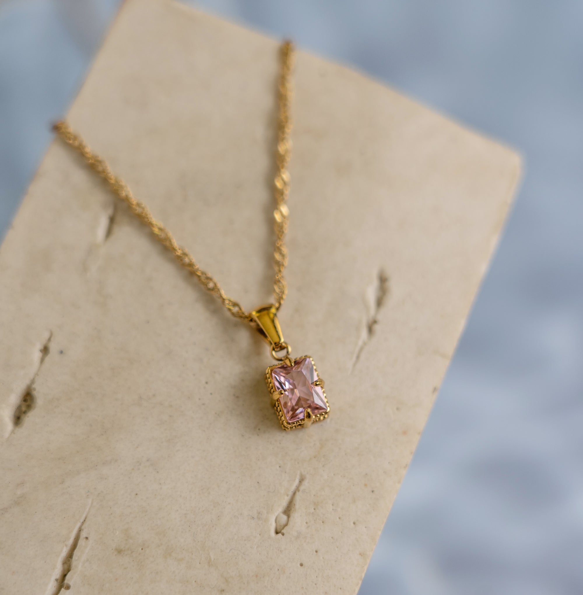 A rose quartz zirconia pendant necklace with a golden chain displayed on a beige card.