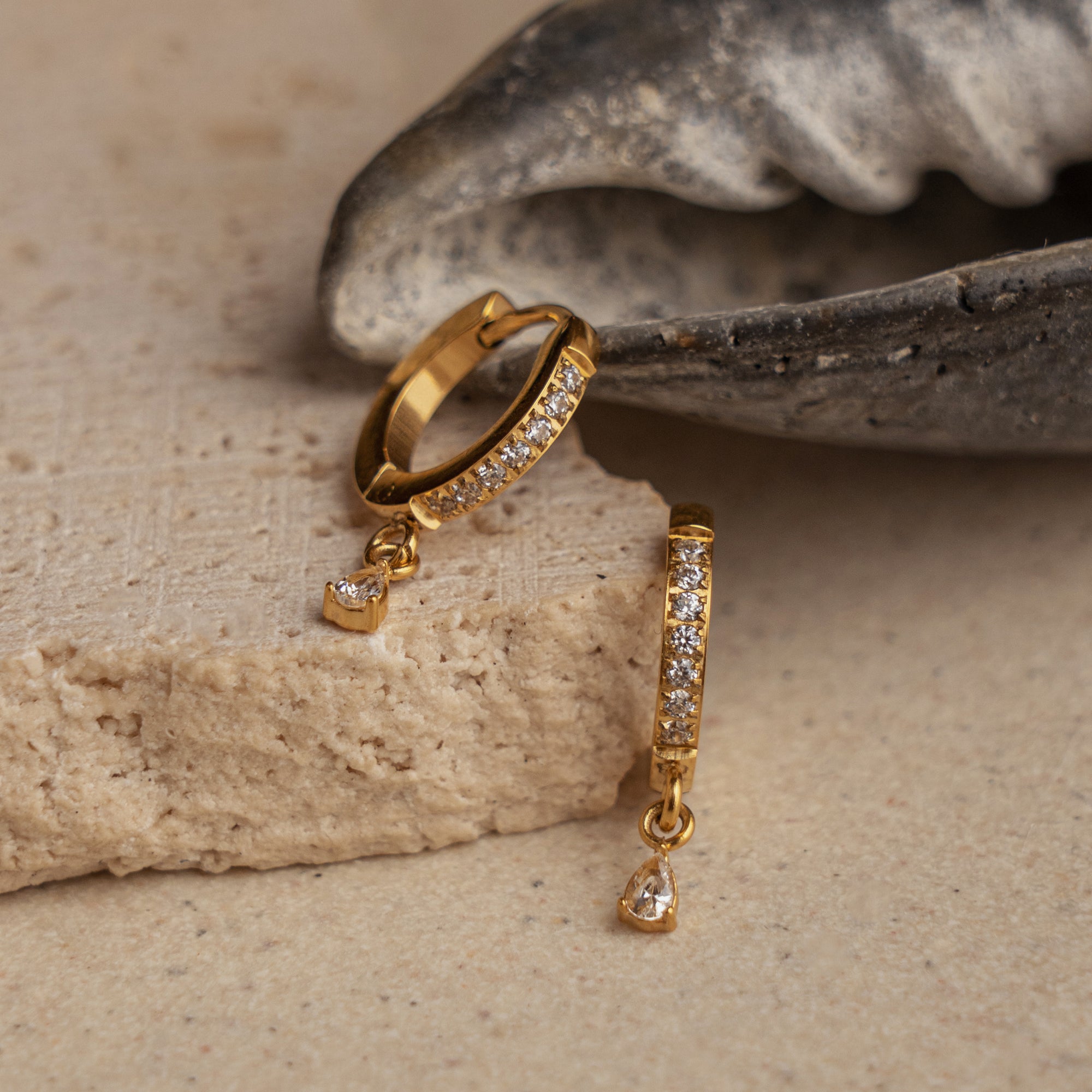 A pair of gold Huggie earrings with a sparkling cubic zirconia charm, displayed on a textured surface.