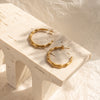 A pair of gold-colored hoop earrings placed on a white wooden surface with a textured background.