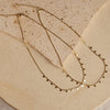A gold-colored necklace with small, regular intervals of pearls displayed on a beige stone surface.